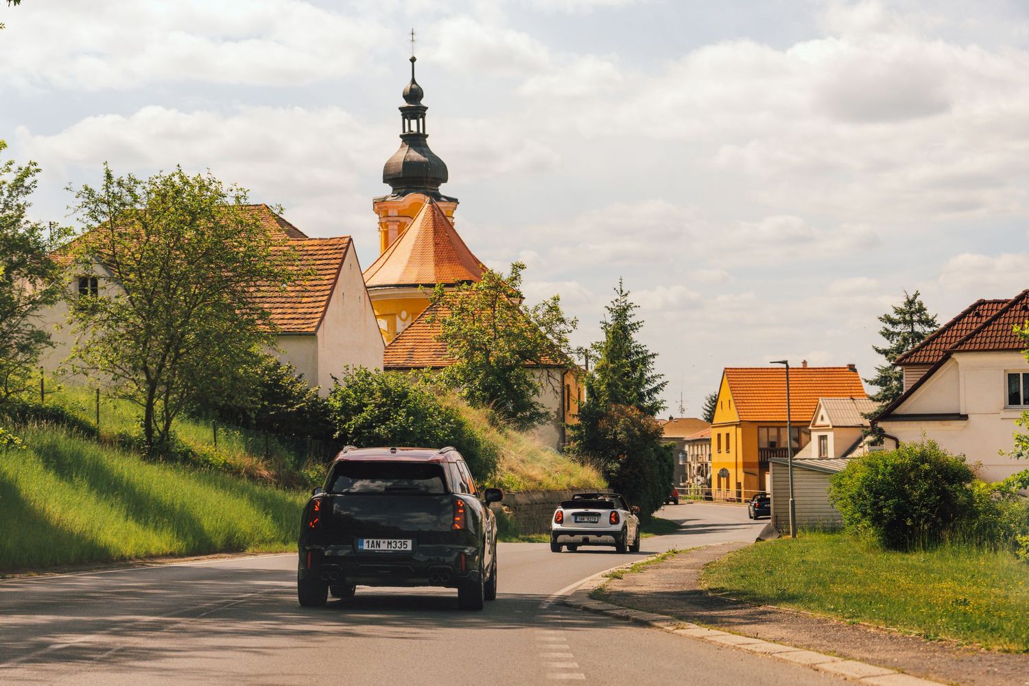 Cars driving through a quaint Czech village road with a historic church tower and colorful houses.