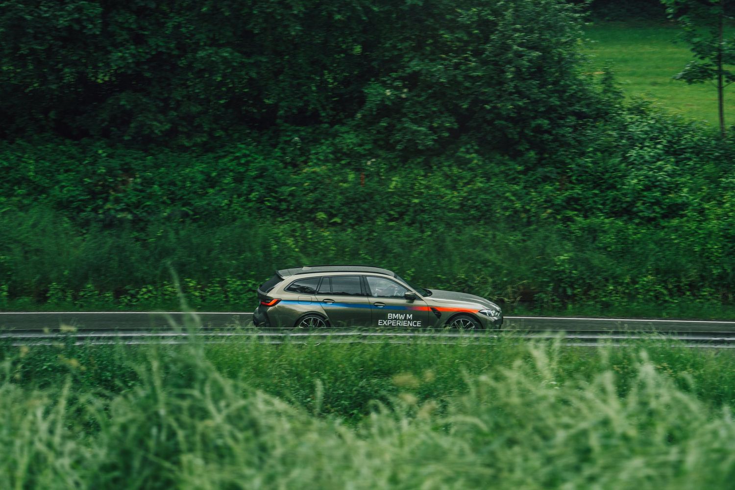 A BMW M3 Touring with BMW Experience livery driving on a road lined with green trees and fields.