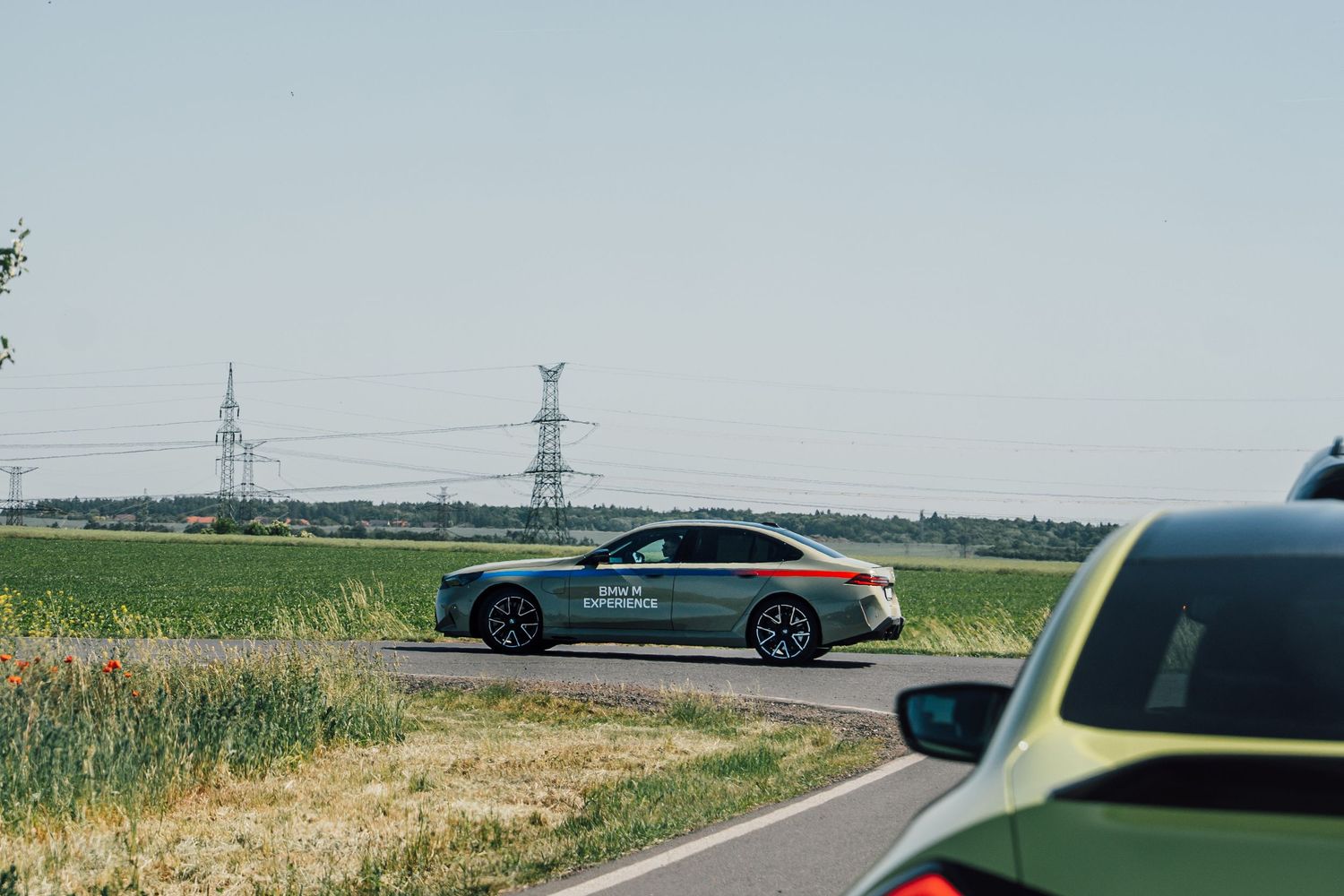 BMW M Experience branded sedan driving on a straight road past green fields and power lines.