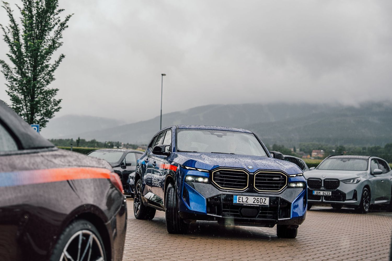 BMW XM in blue camouflage wrap parked with other BMW models against a misty mountain backdrop.