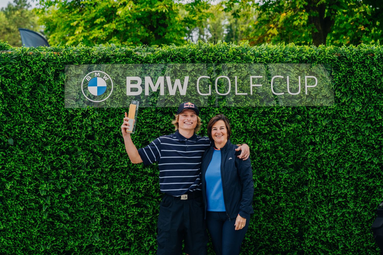Two smiling participants posing in front of a BMW Golf Cup hedge sign at a golf tournament.