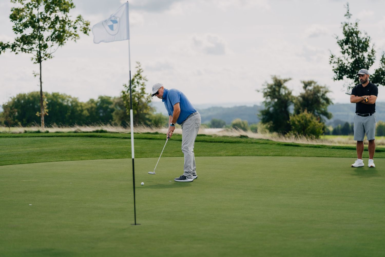 Golfer putting on a green course with a BMW flag, while another player watches in the background.