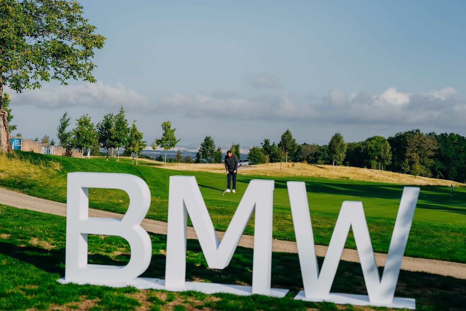 Large white BMW letters sign on a lush green golf course with a person standing behind on a sunny day.