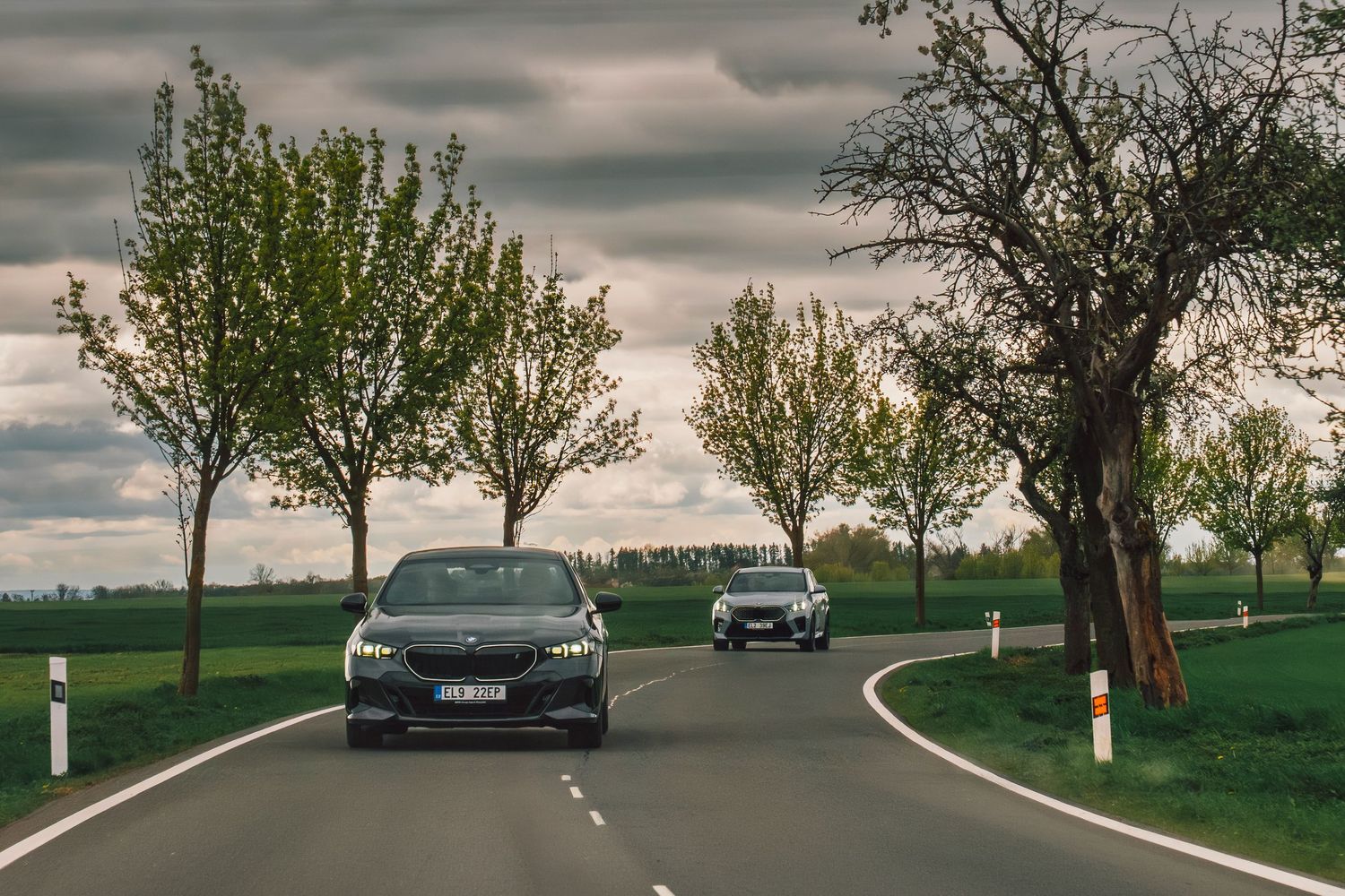 Two BMW sedans driving along a tree-lined country road under moody cloudy skies in spring.