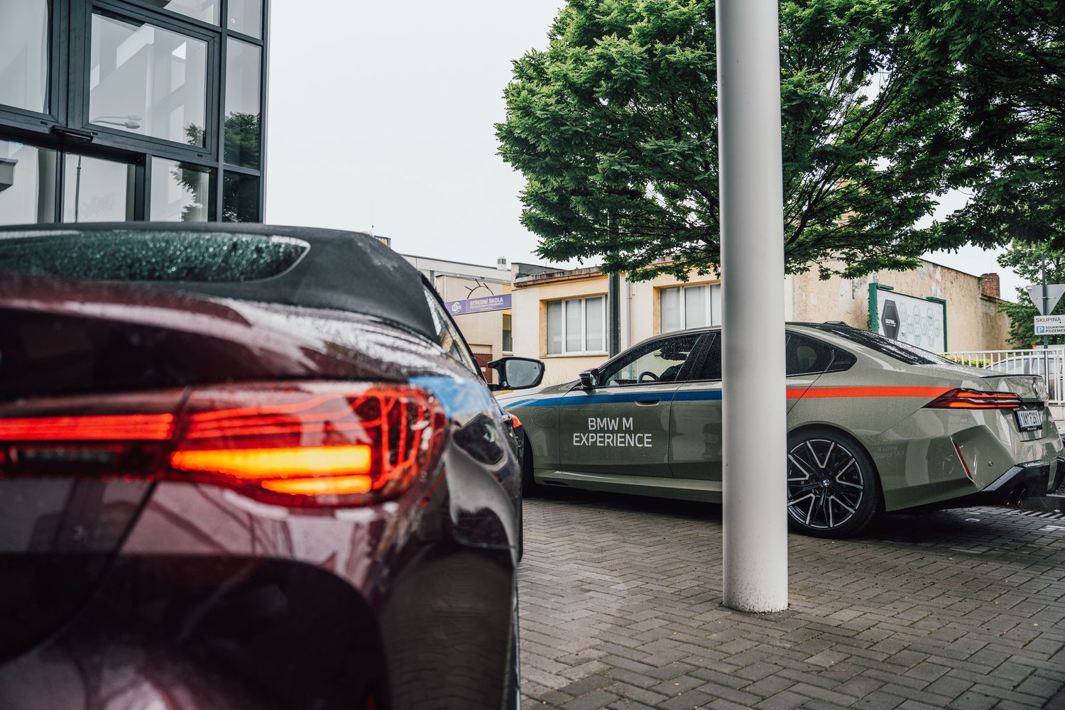 BMW M Experience branded cars parked outside a dealership with lush green trees in background.