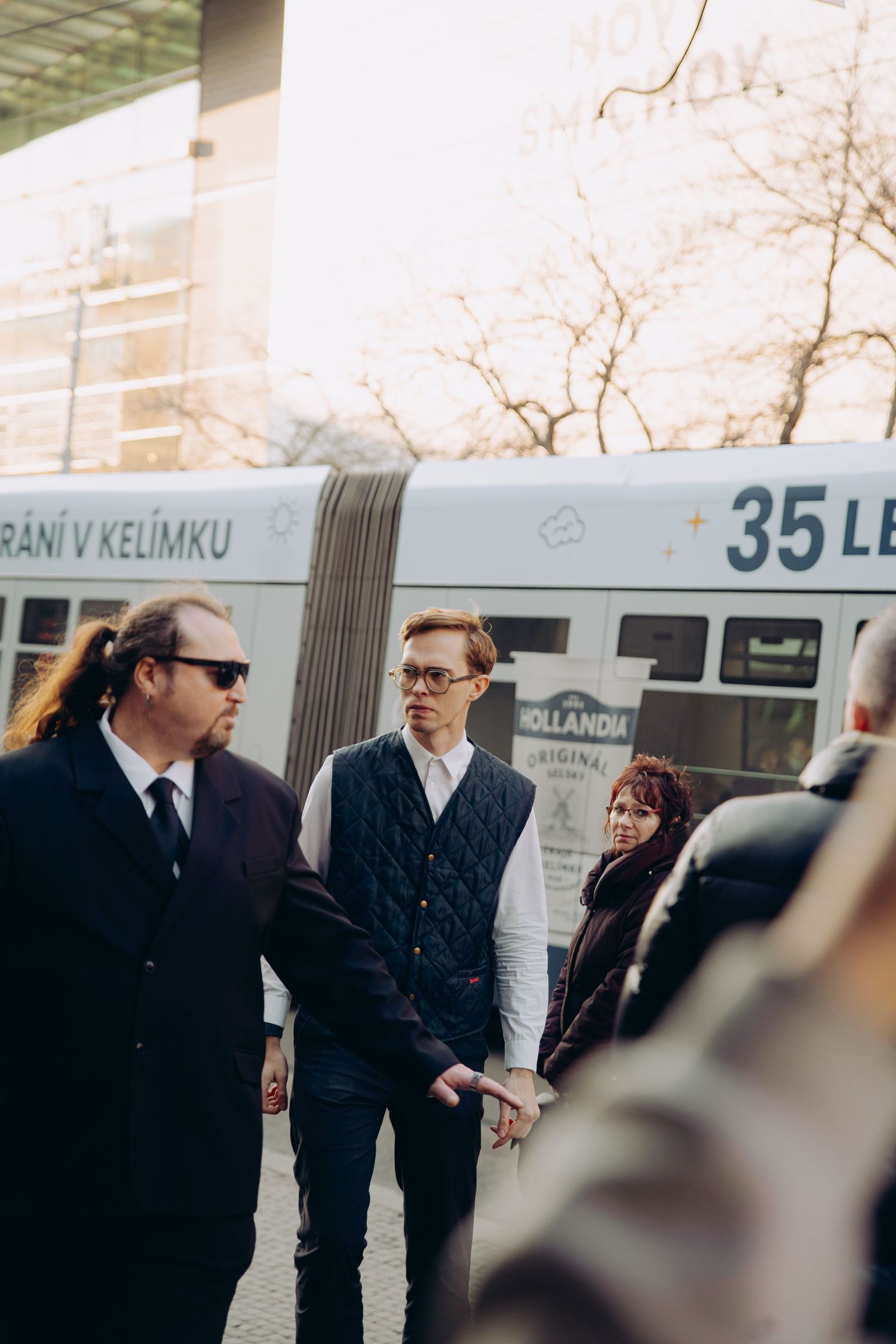 Three people converse near a tram stop on a sunny urban street.