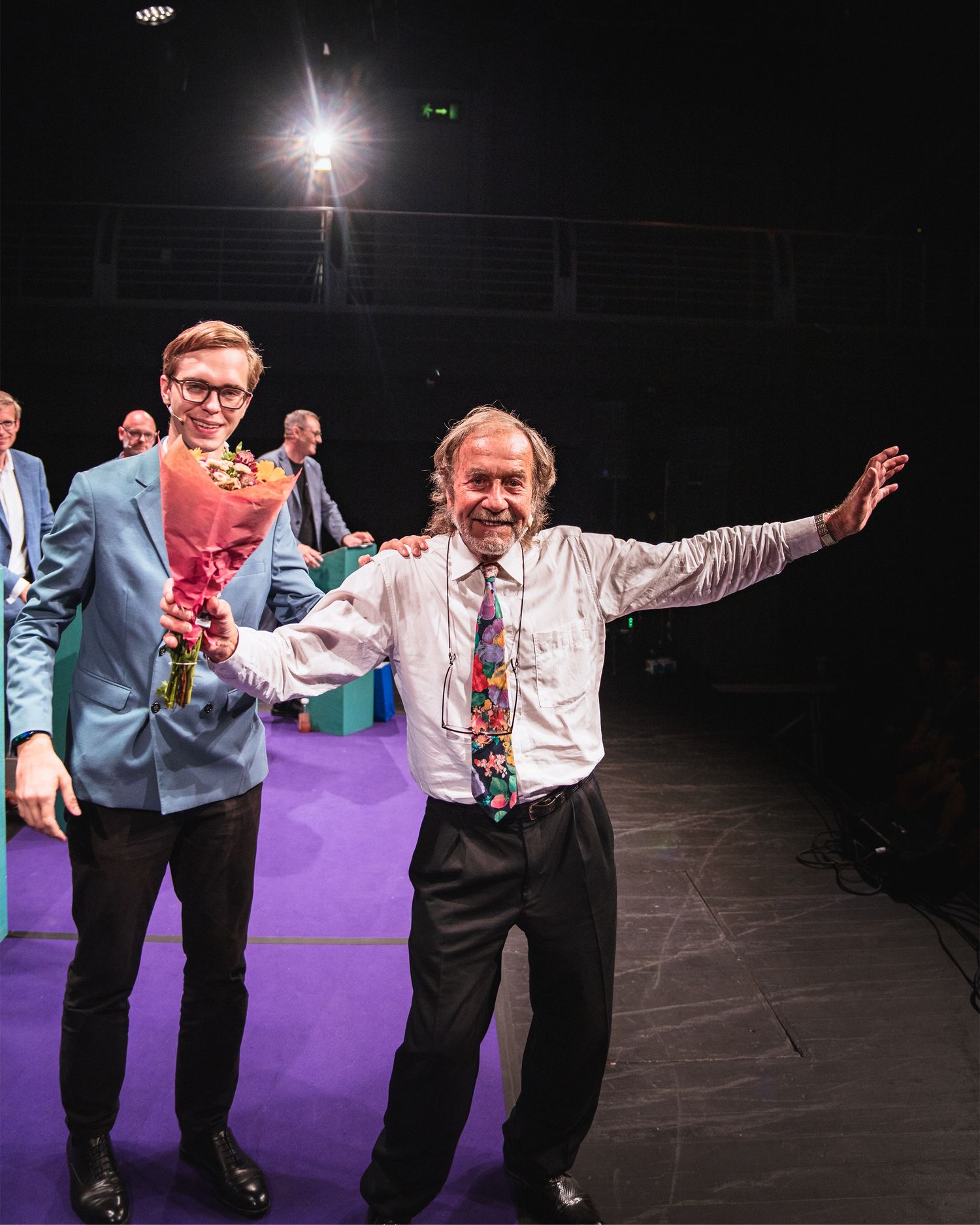 Two men celebrating on stage, one in blue suit holding flowers, other in white shirt with colorful tie.