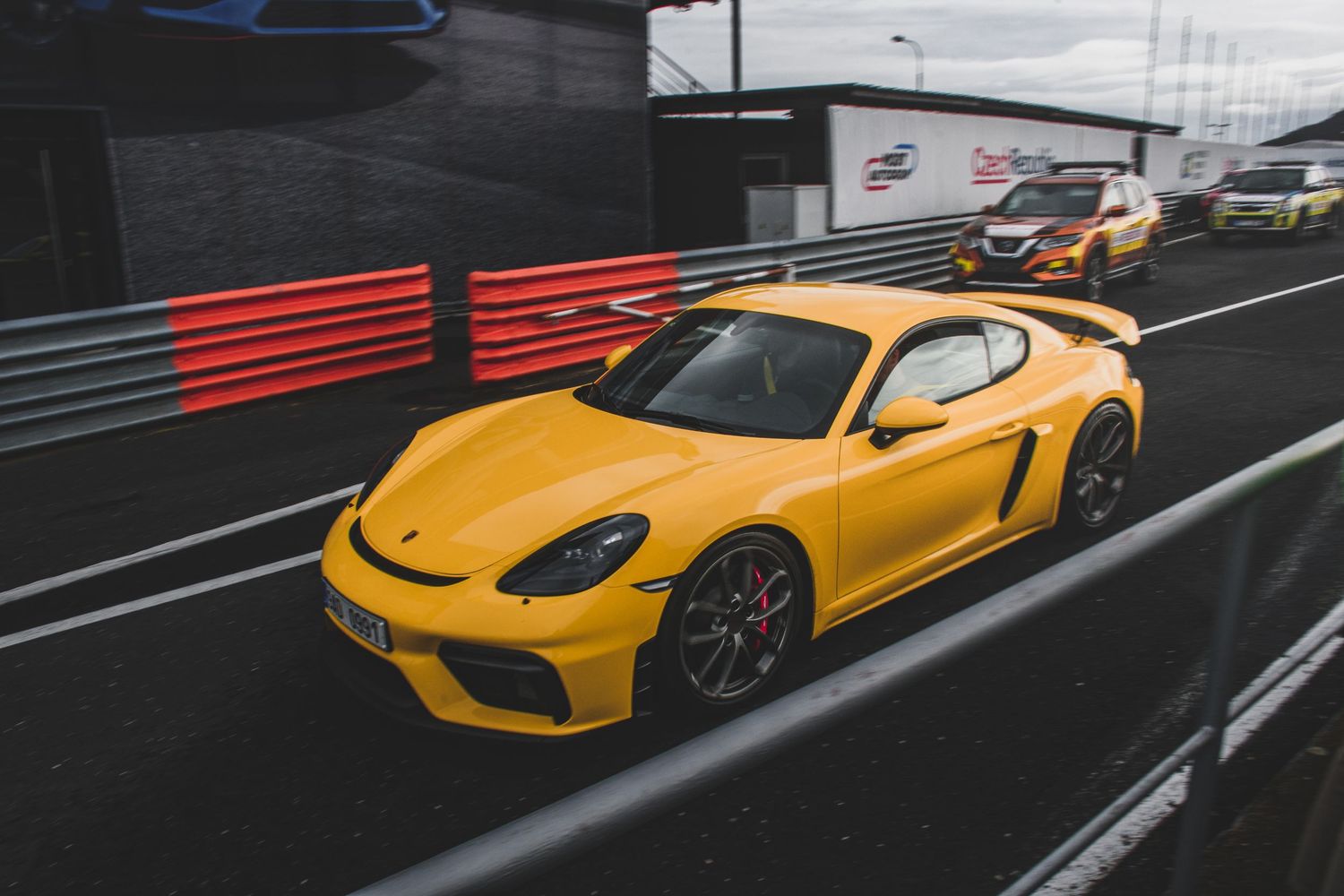 Yellow Porsche 718 Cayman GT4 on a racing circuit with pit lane barriers visible.