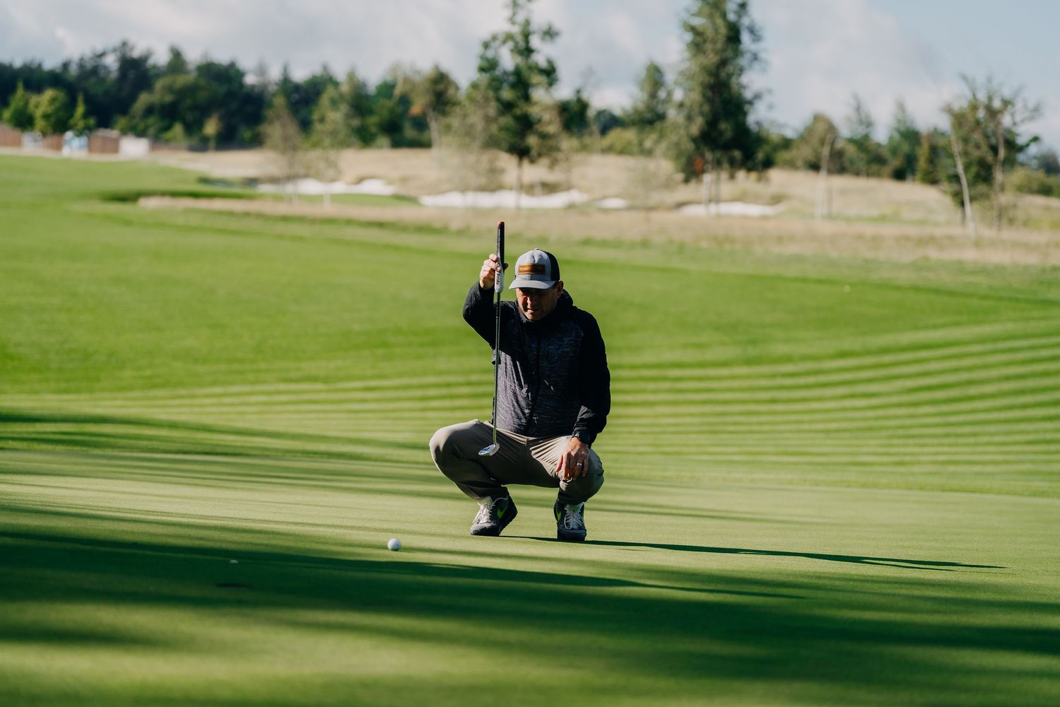 Golfer crouching on green, lining up a putt while holding club upright on a sunny golf course.