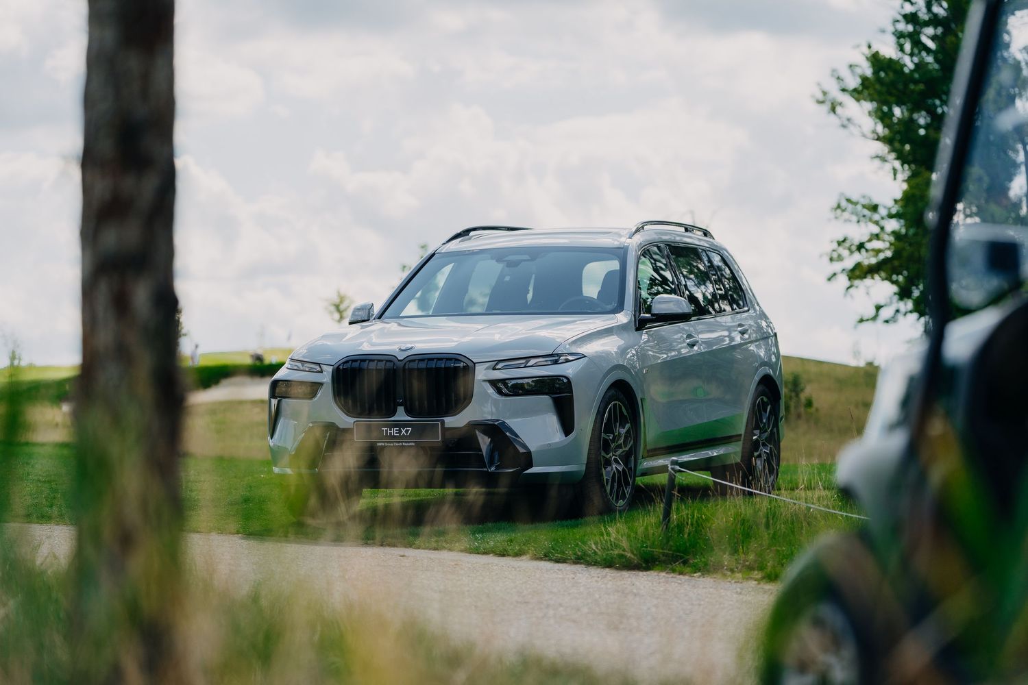 Silver BMW X7 SUV driving on a scenic rural road surrounded by lush green fields and trees.