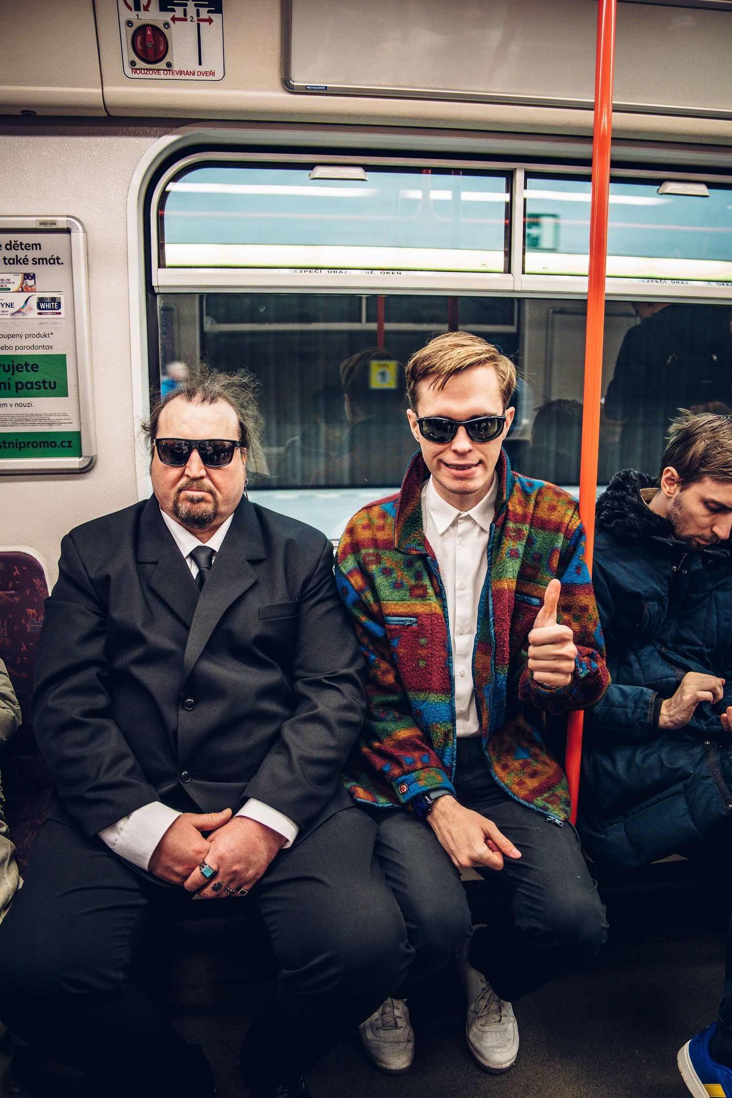 Two men wearing sunglasses sitting on a subway train, one giving a thumbs up in a colorful jacket.