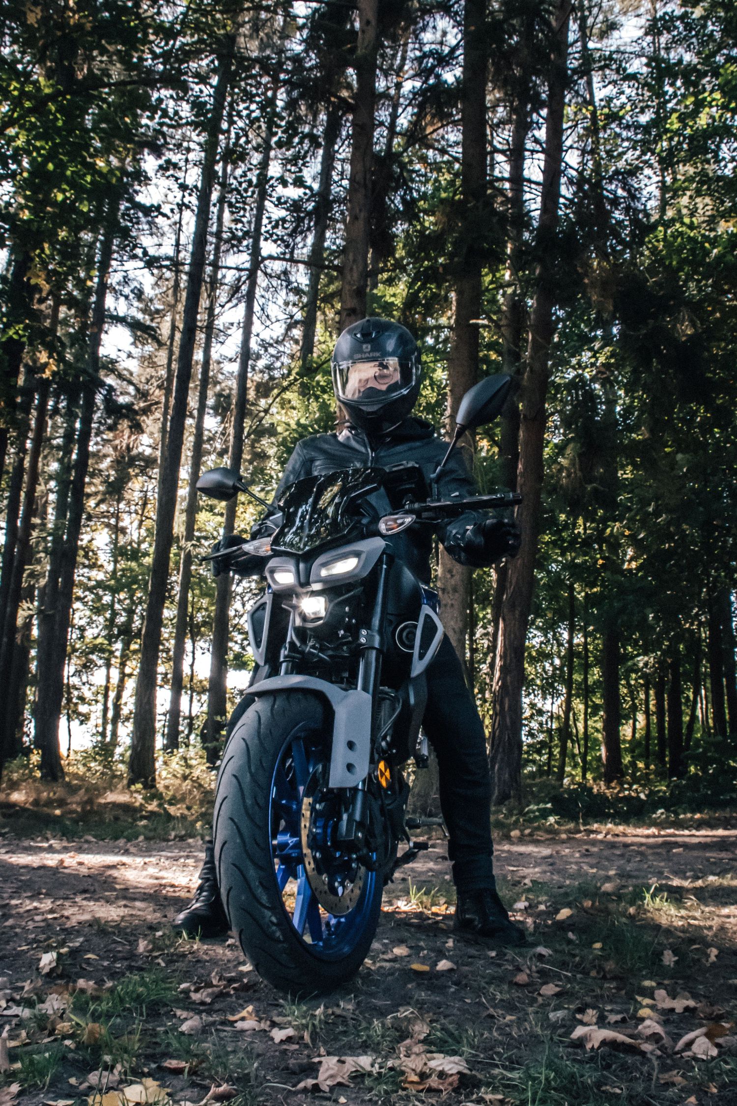 Motorcyclist in black gear and helmet standing beside a blue naked bike in a sunlit forest setting.