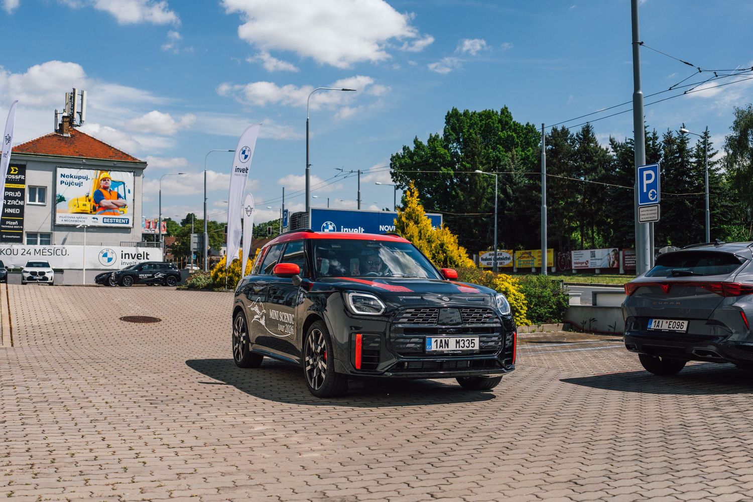 A dark MINI Cooper JCW parked on brick pavement outside a BMW dealership on a sunny day.