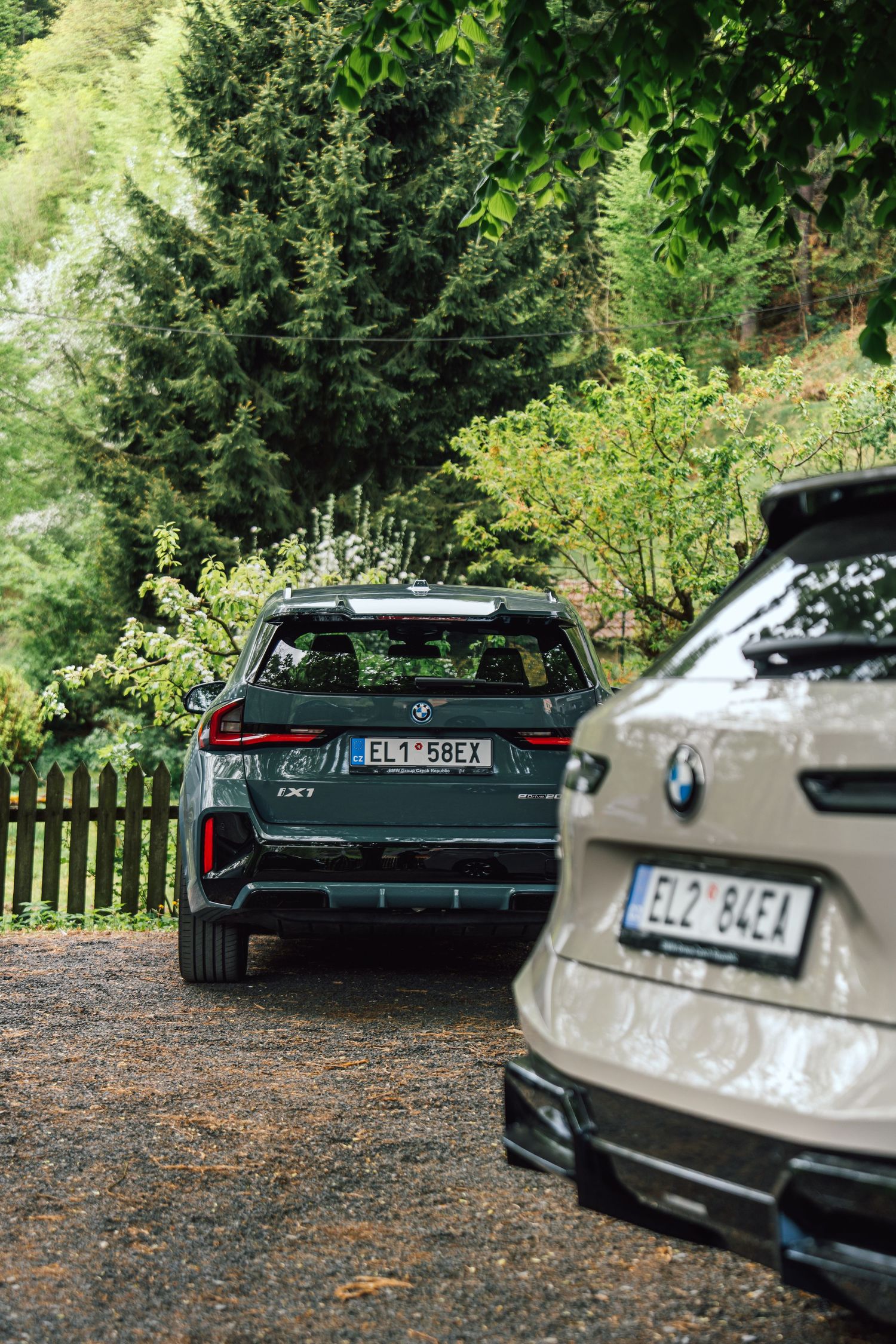 Two BMW electric SUVs parked on a gravel lot surrounded by lush green trees and a wooden fence.