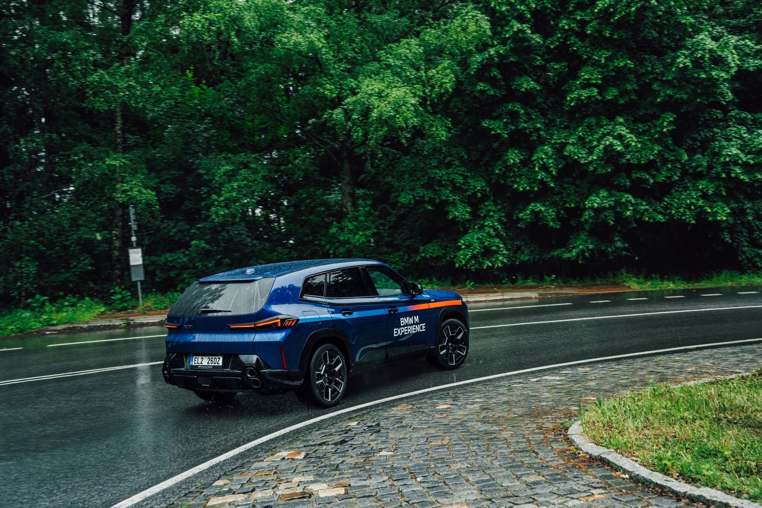 Blue BMW XM driving on a wet road surrounded by lush green forest in the rain.