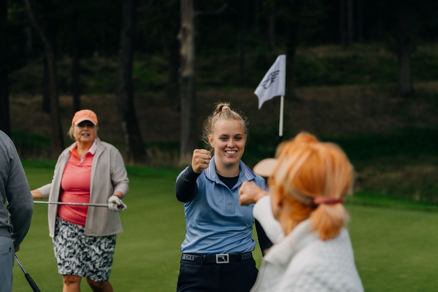 Young woman celebrating on a golf course with fellow players, pumping her fist in victory near the flag.