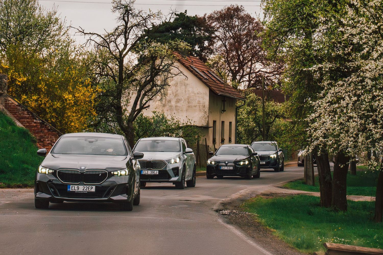 A convoy of BMW electric vehicles driving through a small village surrounded by blooming spring trees.