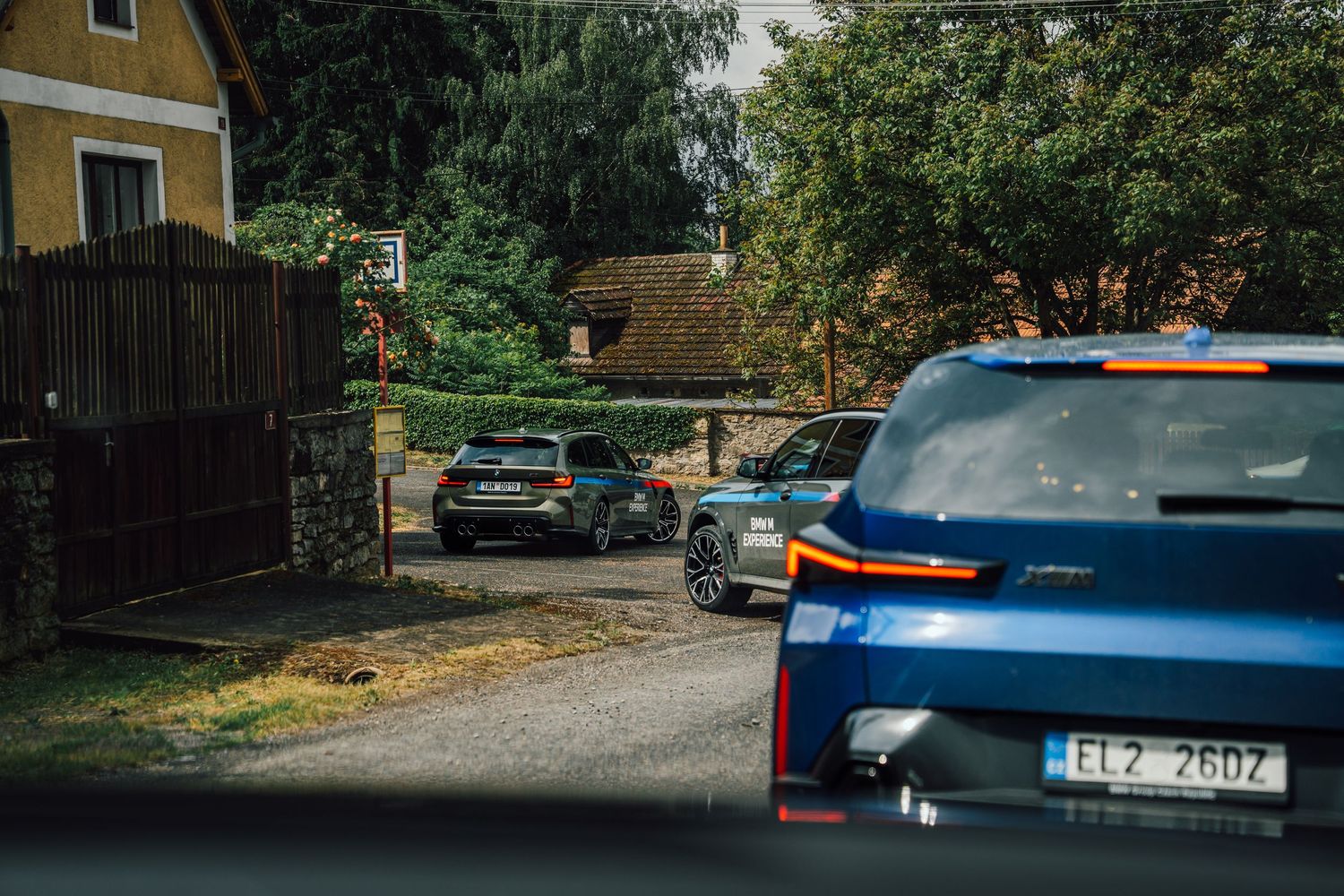 Convoy of BMW M Experience vehicles driving through a narrow rural Czech village road.