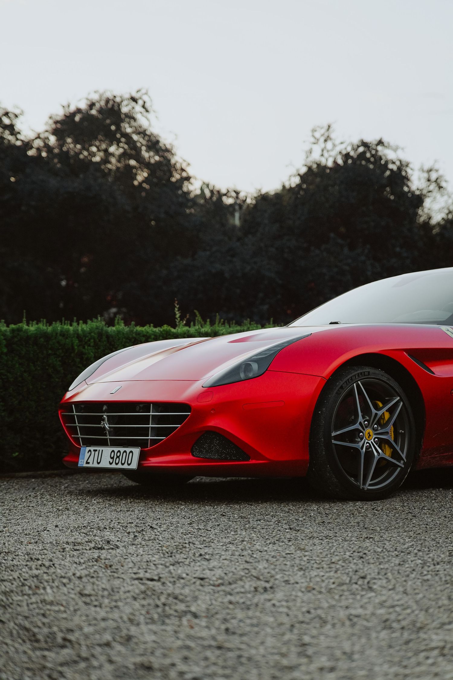 Red Ferrari California T parked on gravel with Czech license plate, surrounded by lush green hedges at dusk.