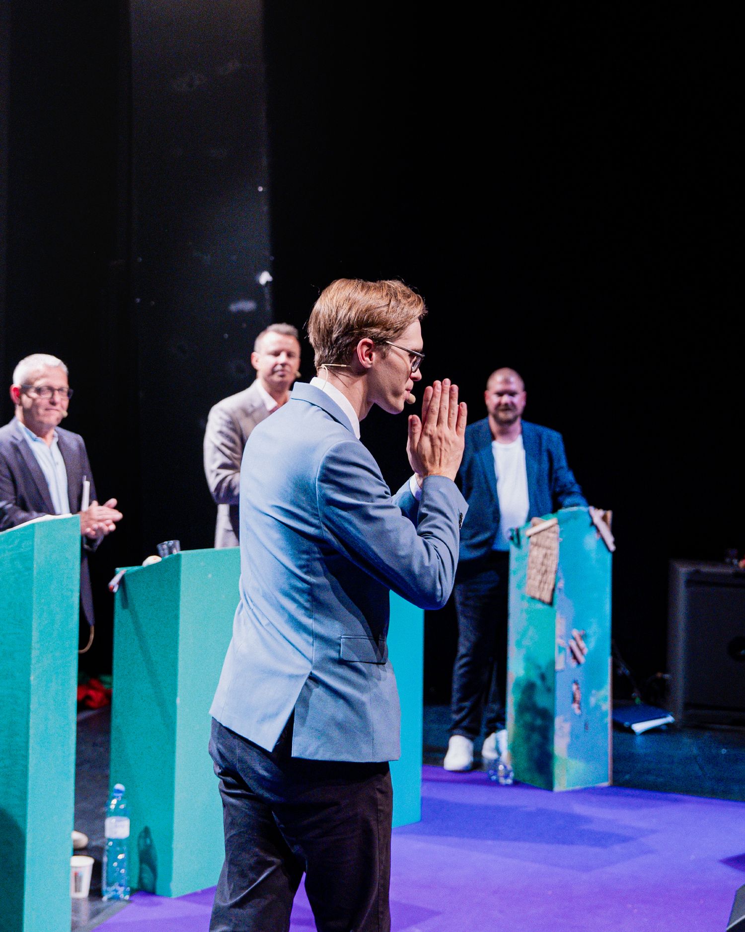 Young man in light blue suit bowing with hands pressed together on stage before audience.