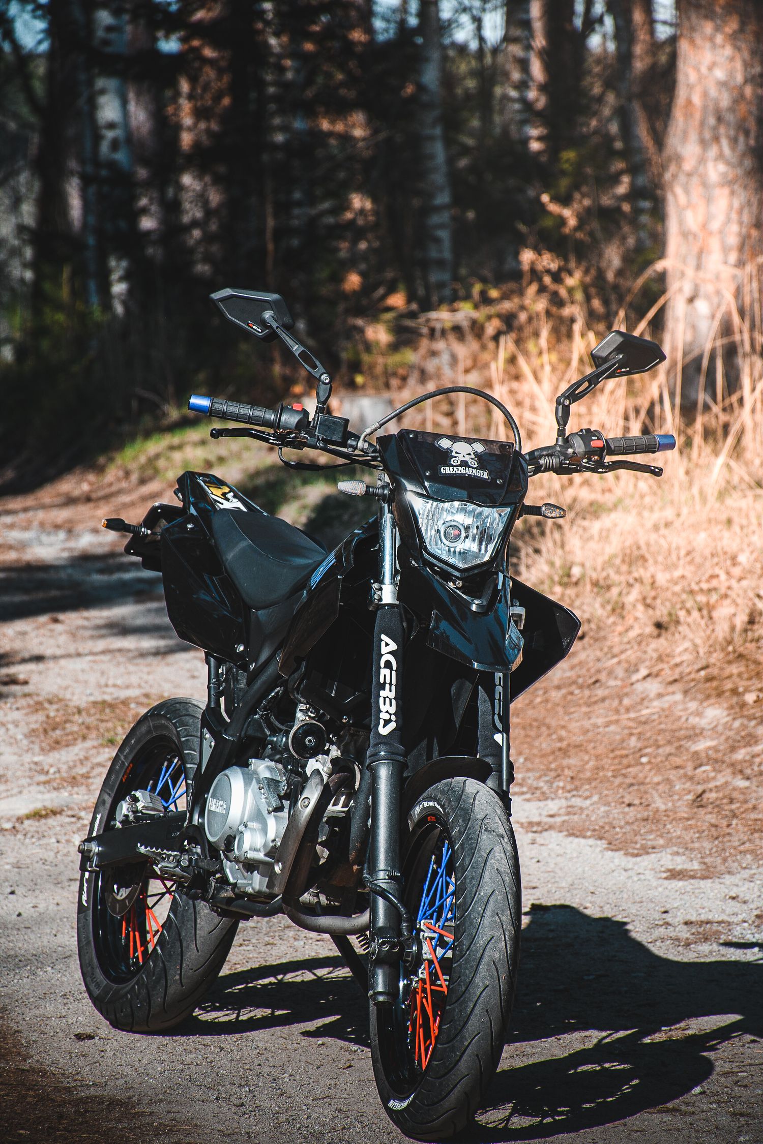 Black Acerbis supermoto motorcycle parked on a sunlit dirt road surrounded by trees.