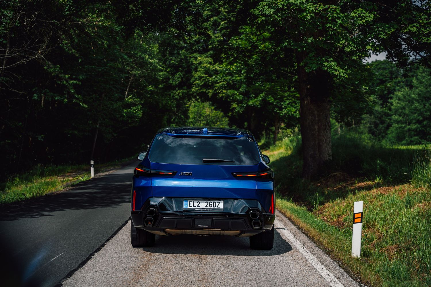 Rear view of a blue BMW iX driving on a tree-lined road in lush green forest surroundings.