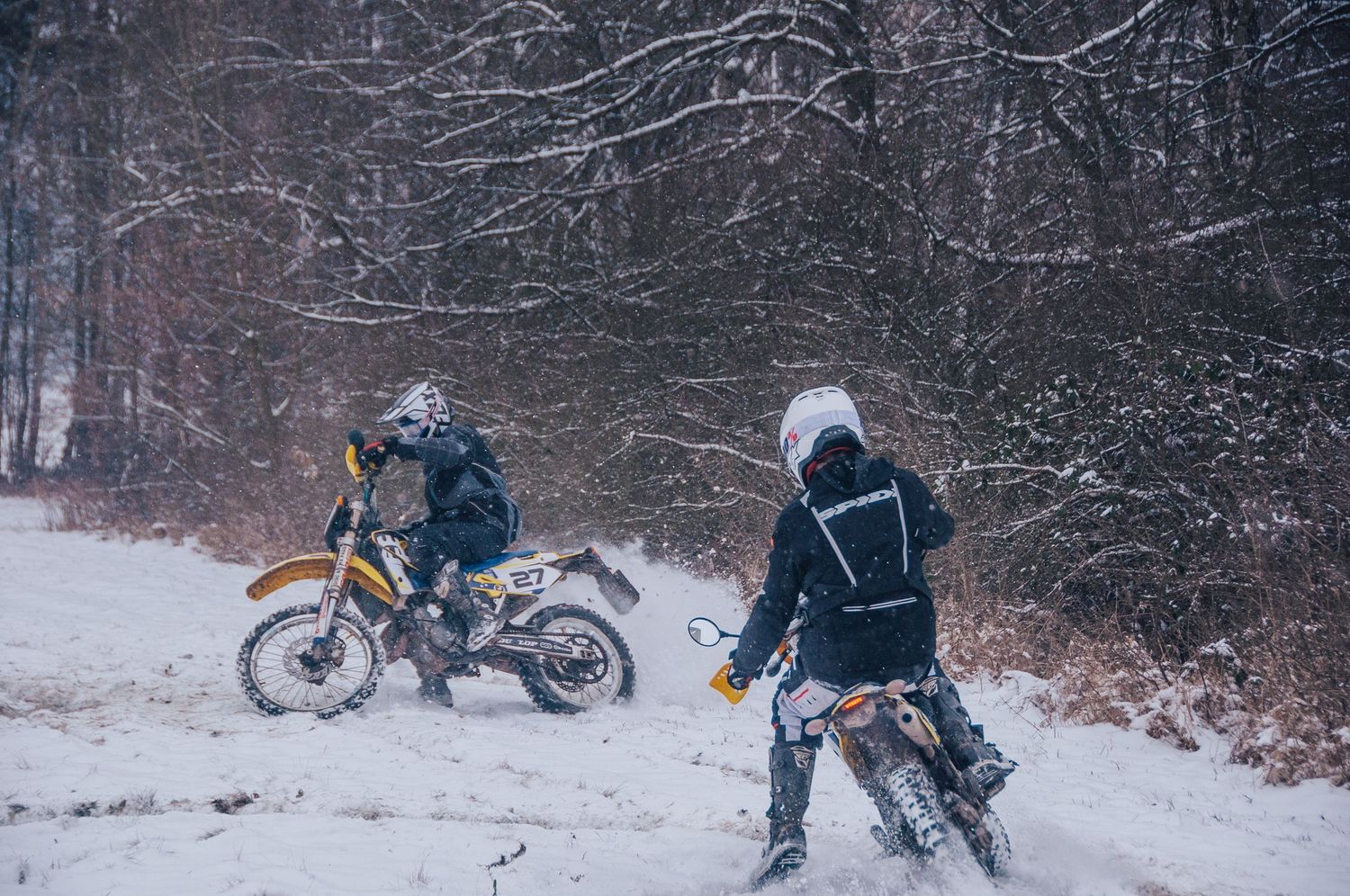 Two motocross riders navigate snowy forest trail on dirt bikes during winter snowfall.