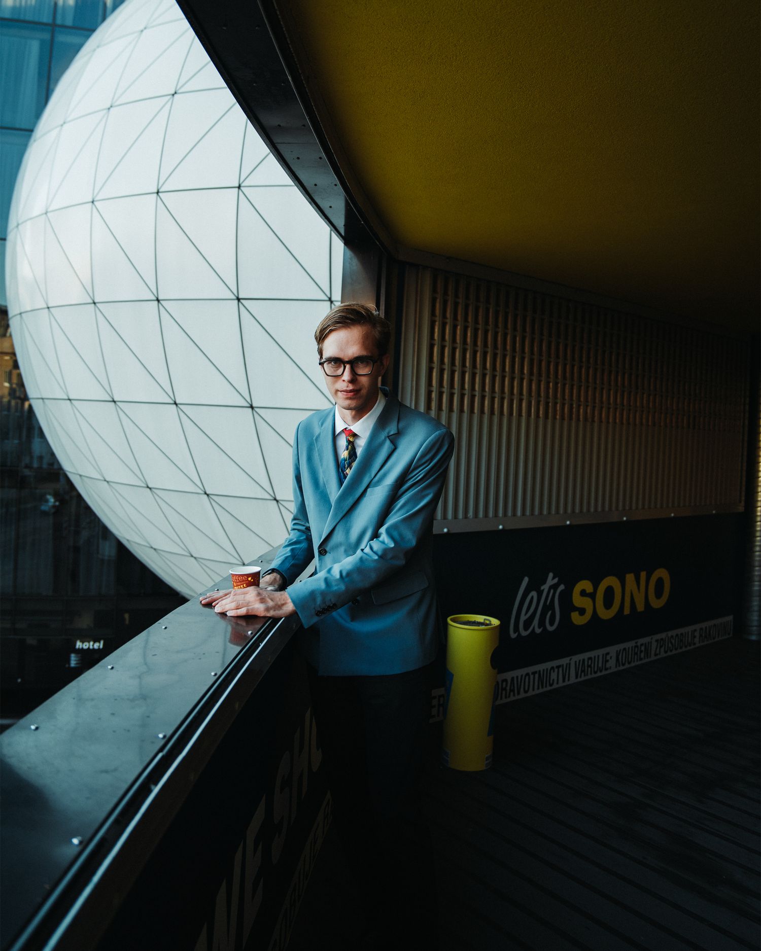 Stylish man in blue blazer and glasses stands on a balcony at Sono venue.