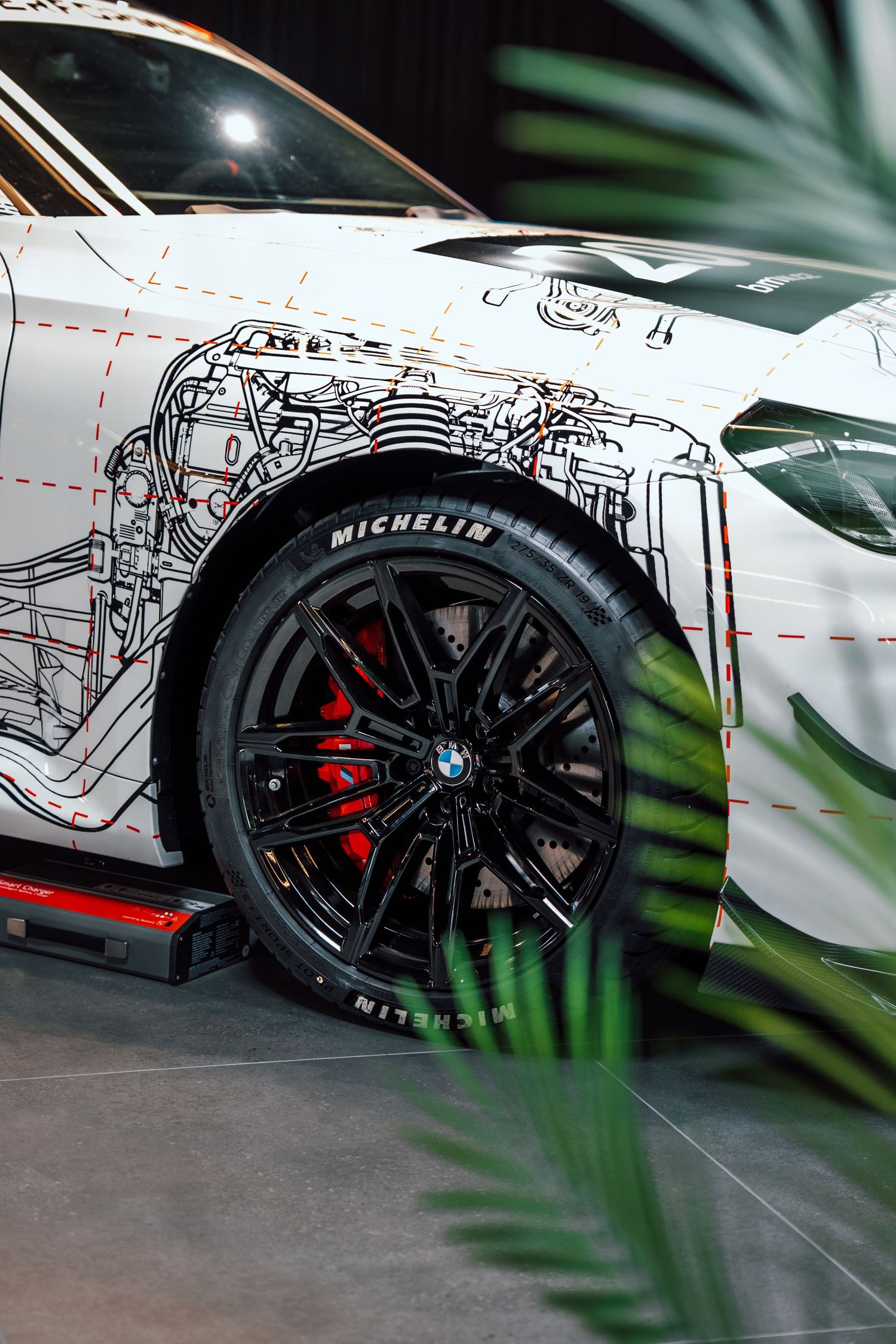 Close-up of a BMW M race car wheel with Michelin tire and red brake calipers, featuring technical blueprint artwork.