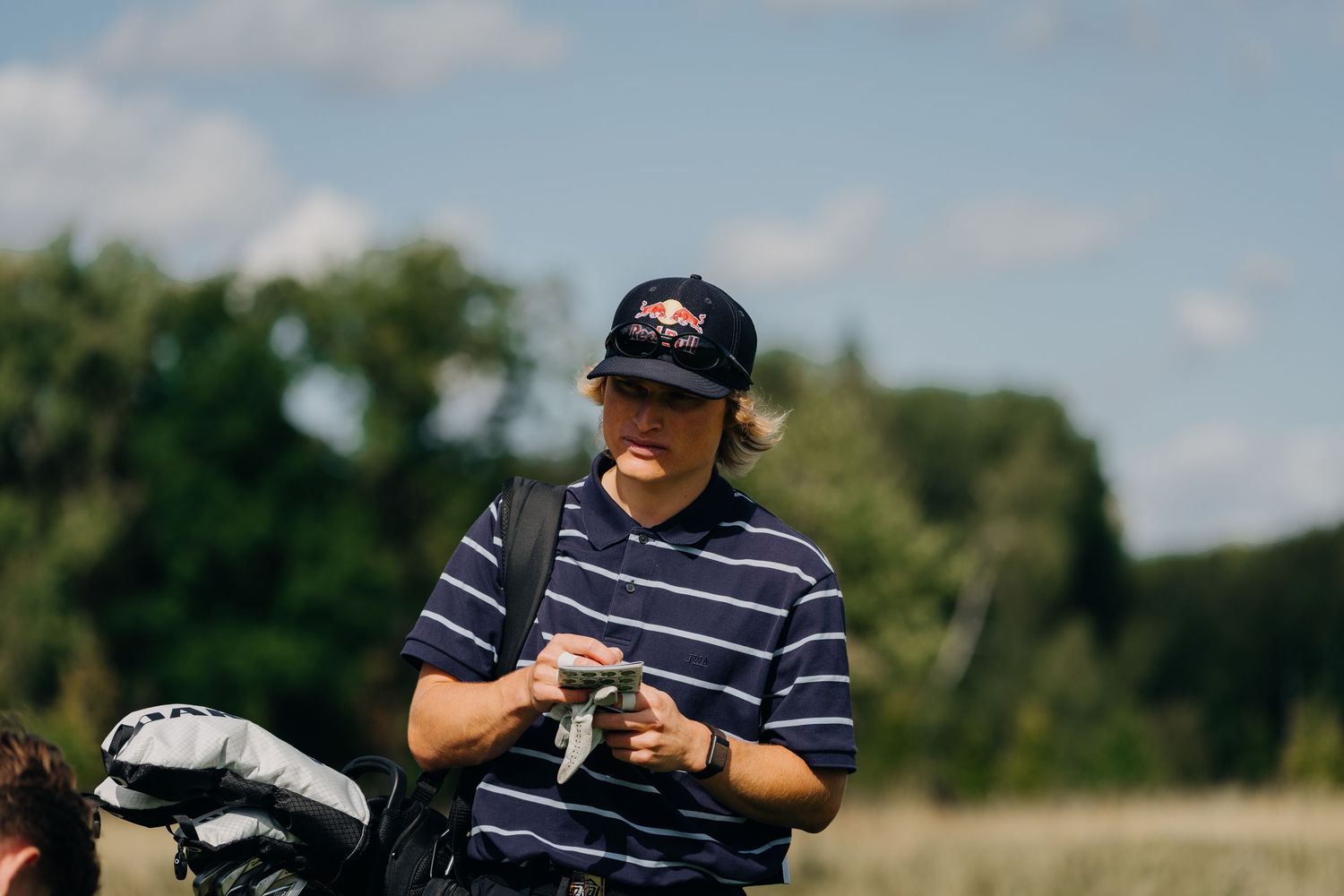 Golfer wearing a Red Bull cap and striped polo shirt reviewing scorecard on a sunny golf course.