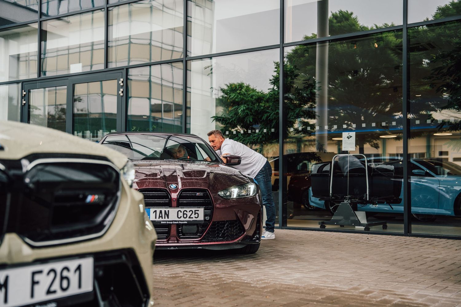 Man examining a dark burgundy BMW M3 at a BMW dealership showroom with glass facade.