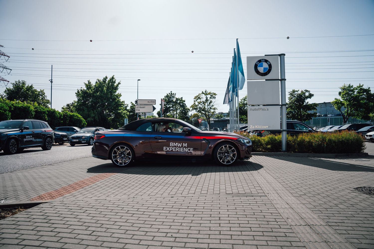 BMW M Experience car passing a BMW dealership sign on a sunny urban street.