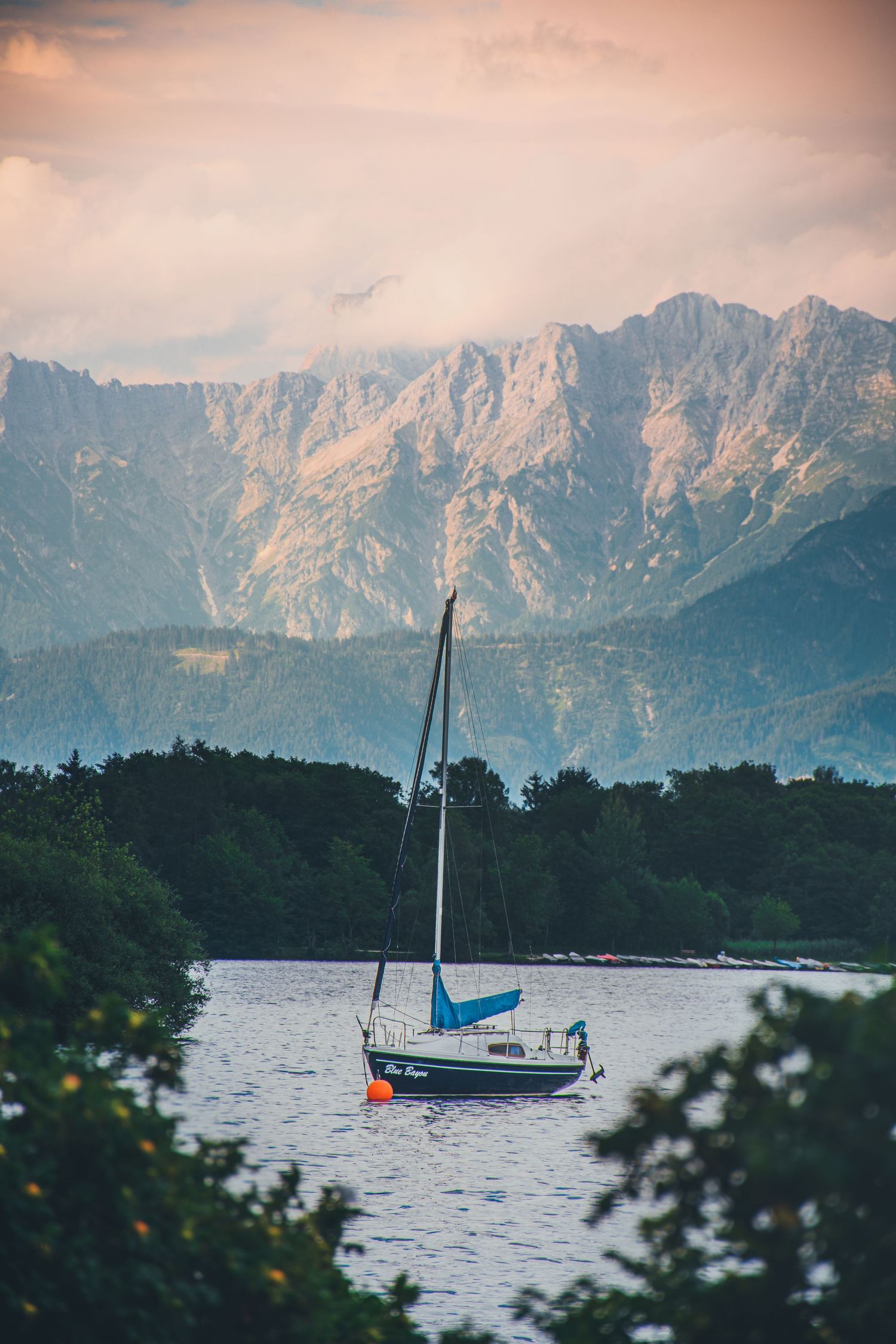 A sailboat anchored on a calm alpine lake with dramatic mountain peaks and colorful sunset sky in the background.