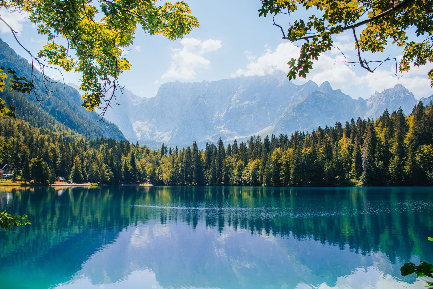 A crystal-clear turquoise alpine lake reflecting pine forests and snow-capped mountain peaks on a sunny day.