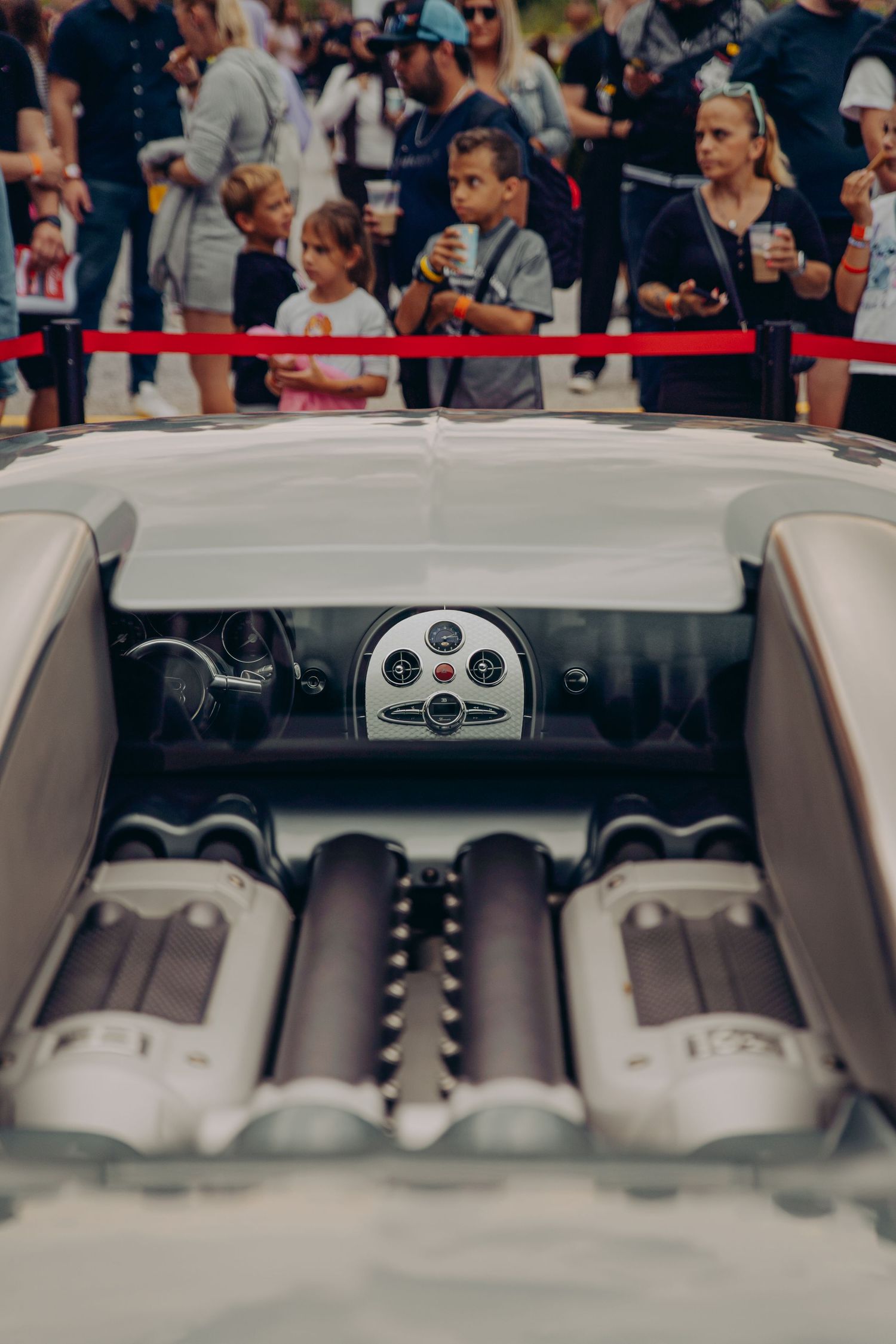 Rear cockpit view of a classic silver open-top sports car with exposed engine cylinders at a car show.