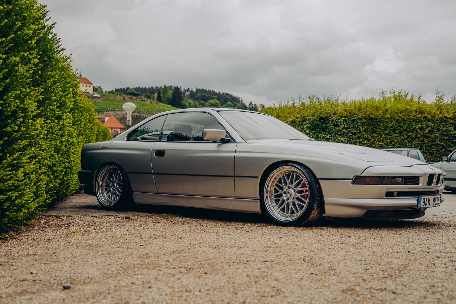 Silver BMW 8 Series E31 coupe with BBS wheels and red brake calipers parked beside a green hedge.