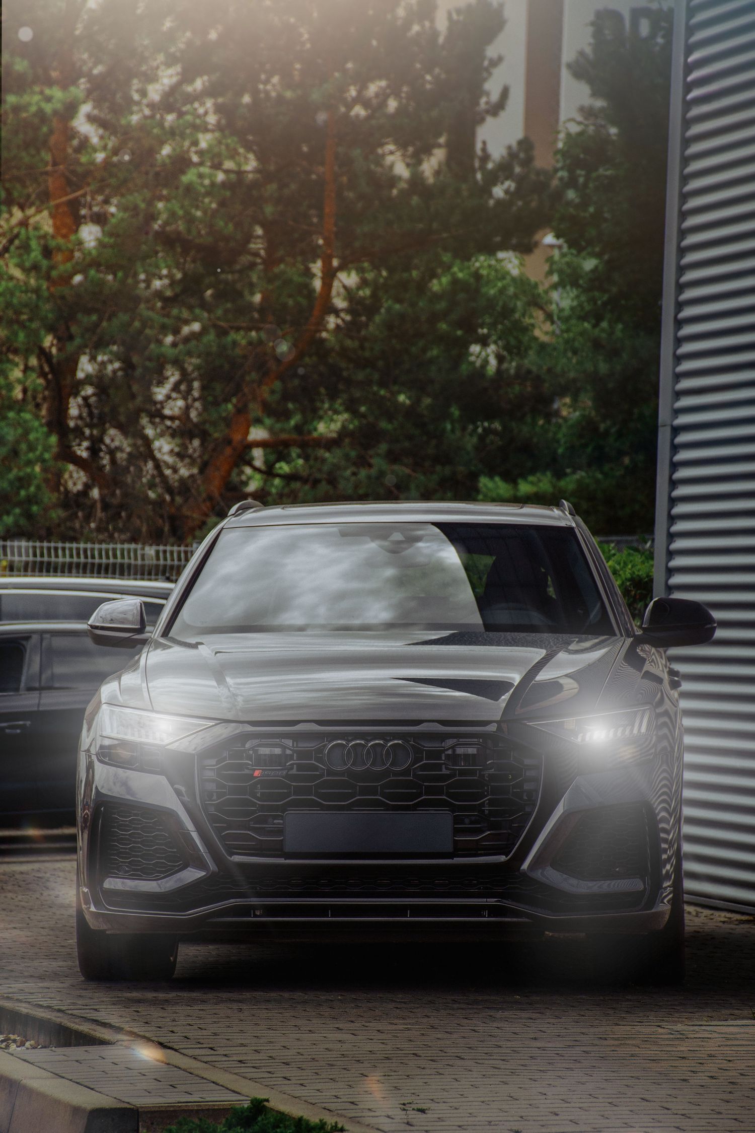 Black Audi SUV with illuminated headlights parked outside a building surrounded by green trees.