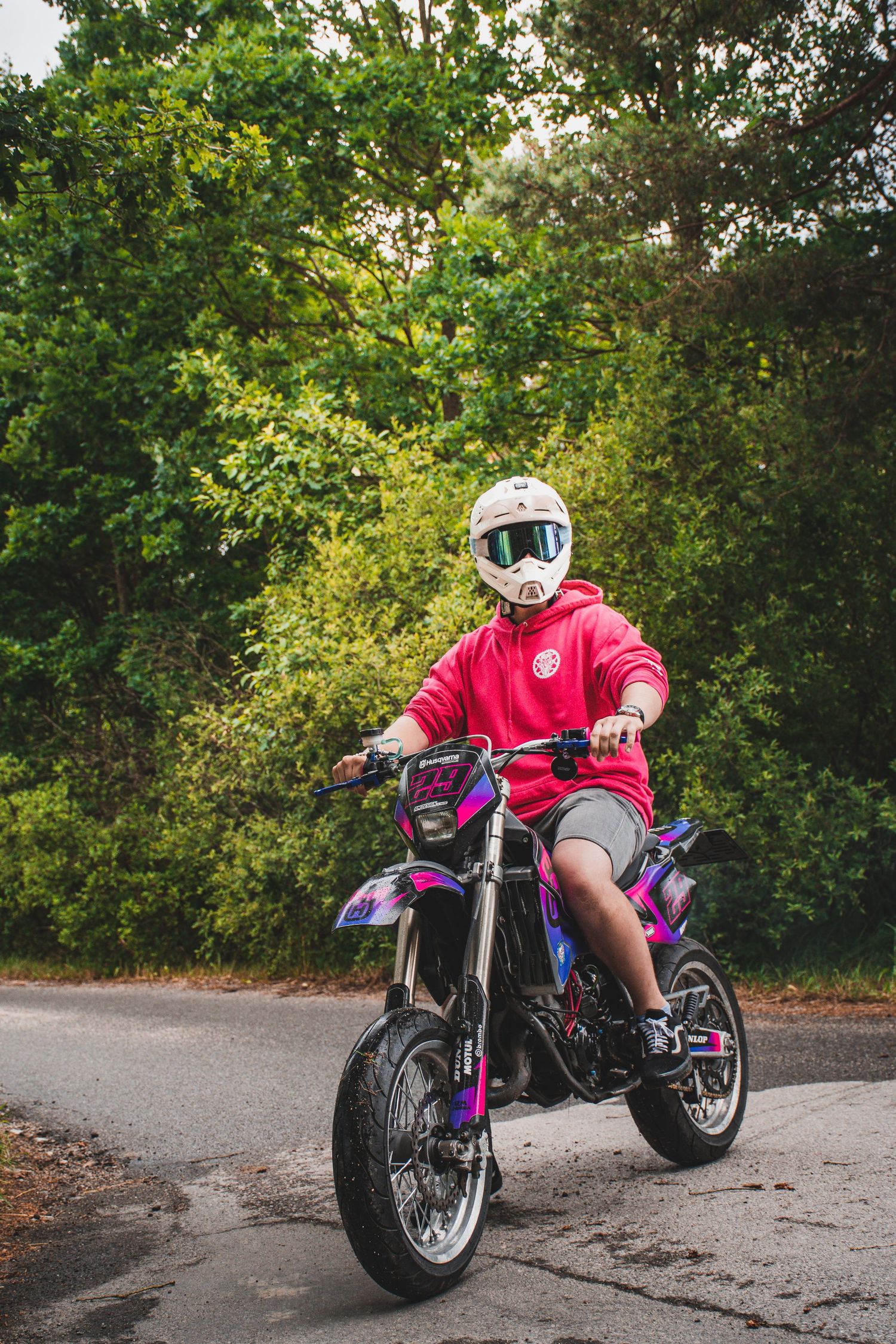 Rider in white helmet and red hoodie riding a purple and pink dirt bike on a road.