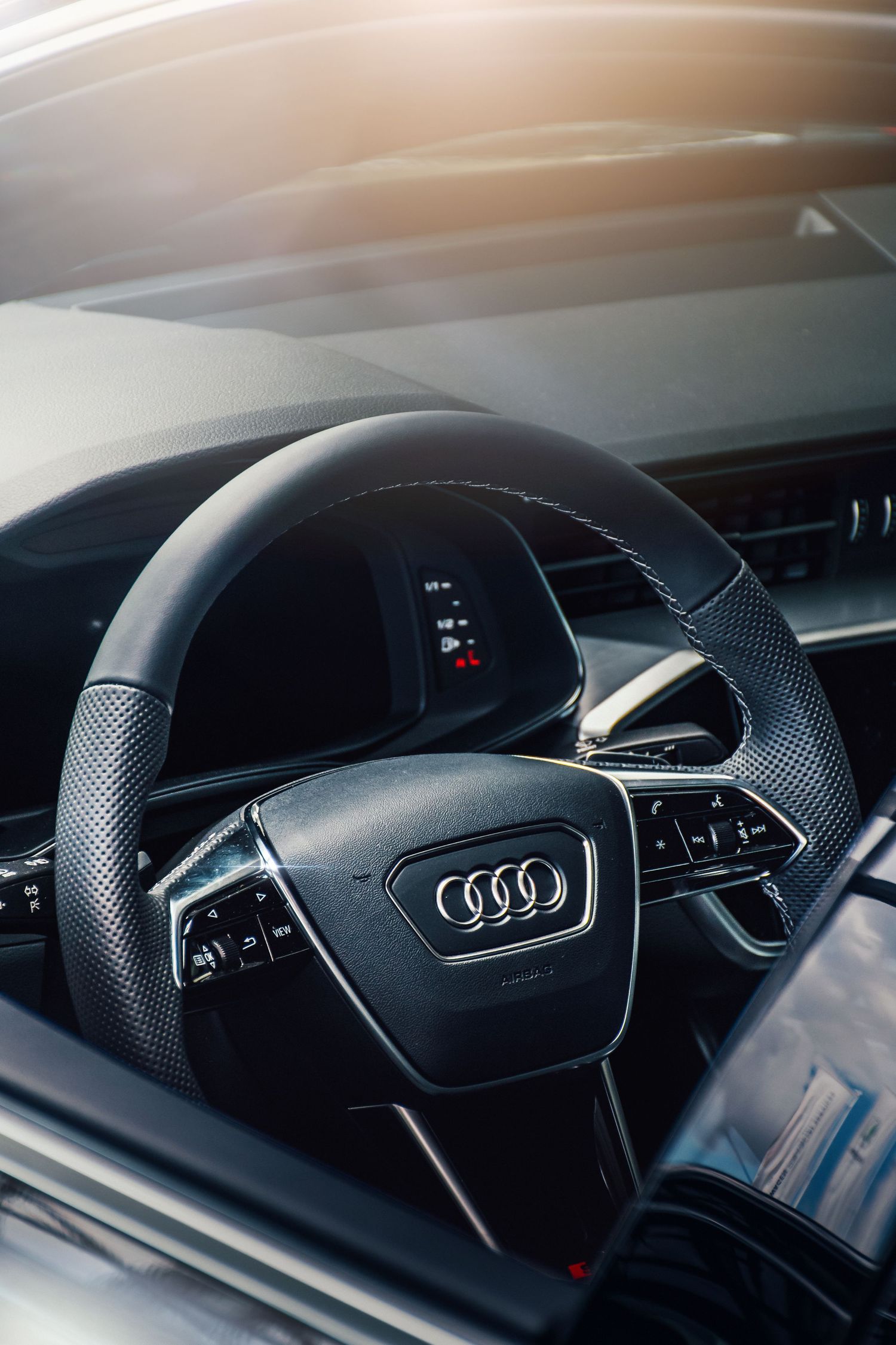 Close-up of a black Audi leather steering wheel with four rings logo illuminated by sunlight in car interior.