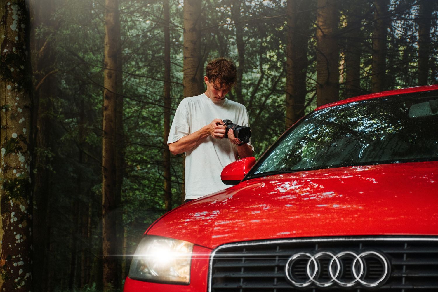 Young man reviewing photos on camera next to a red Audi car in a forest.