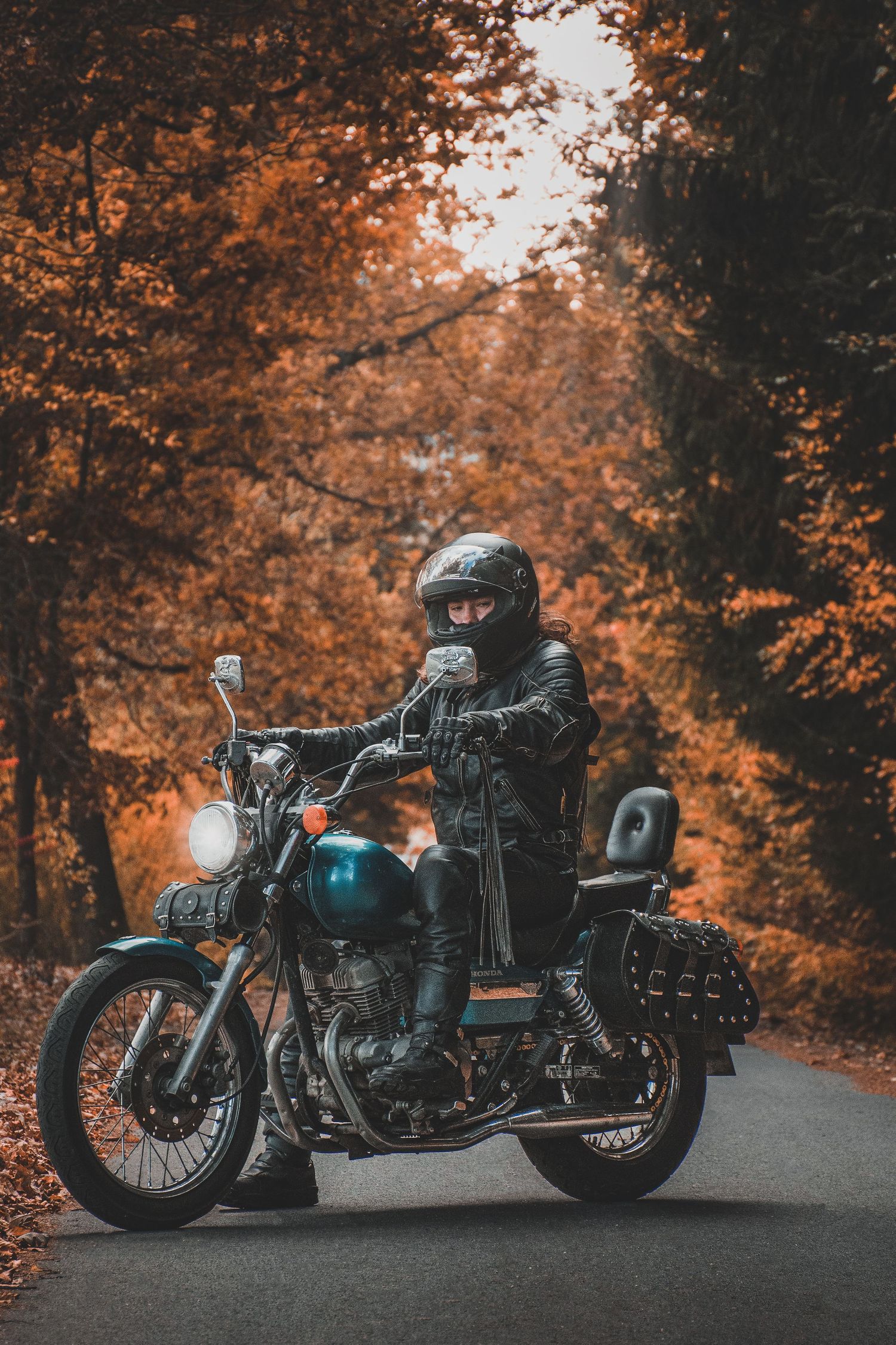 Biker in black leather jacket and helmet posing on classic cruiser motorcycle on autumn forest road.