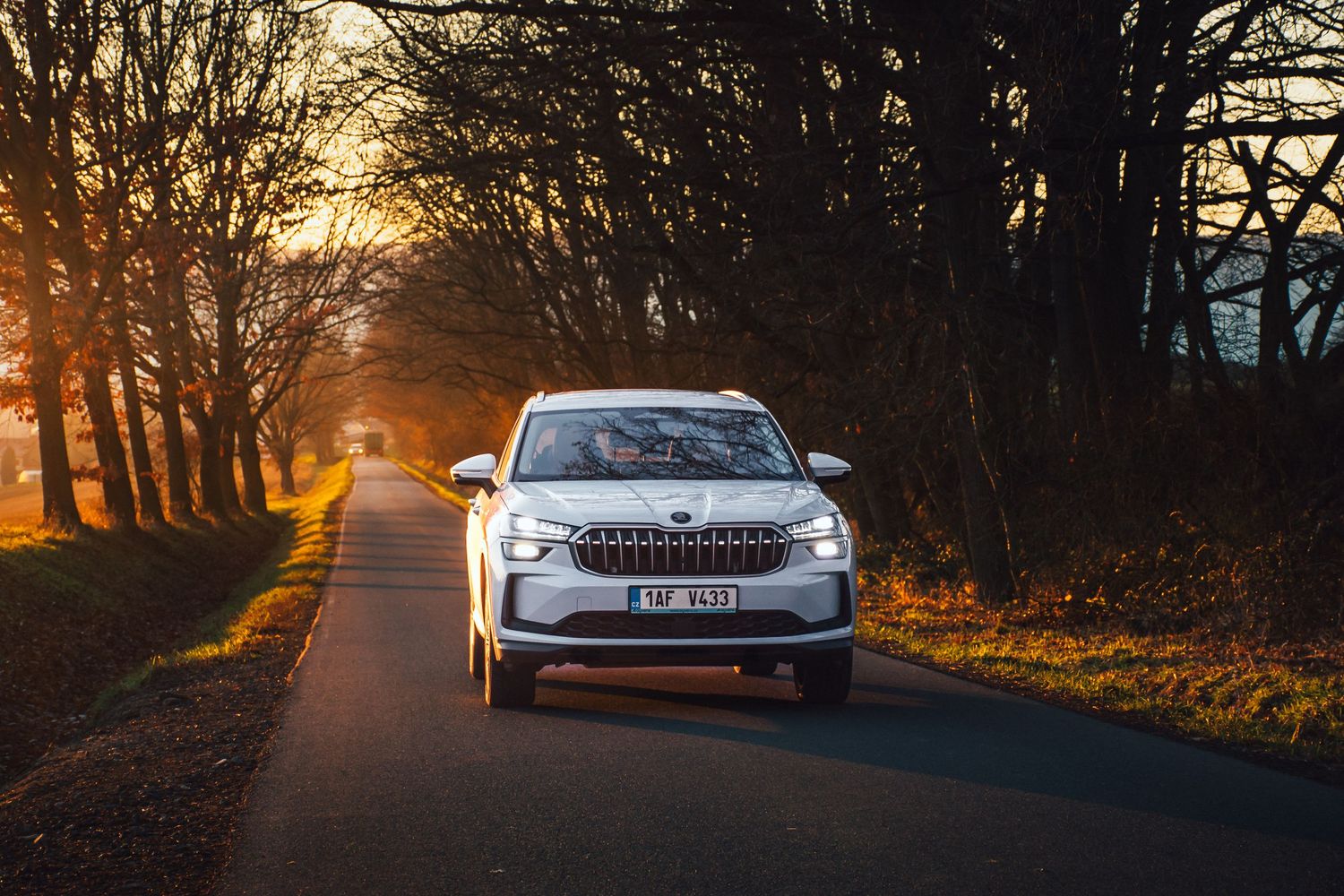 White modern SUV driving on a tree-lined road at golden sunset hour.