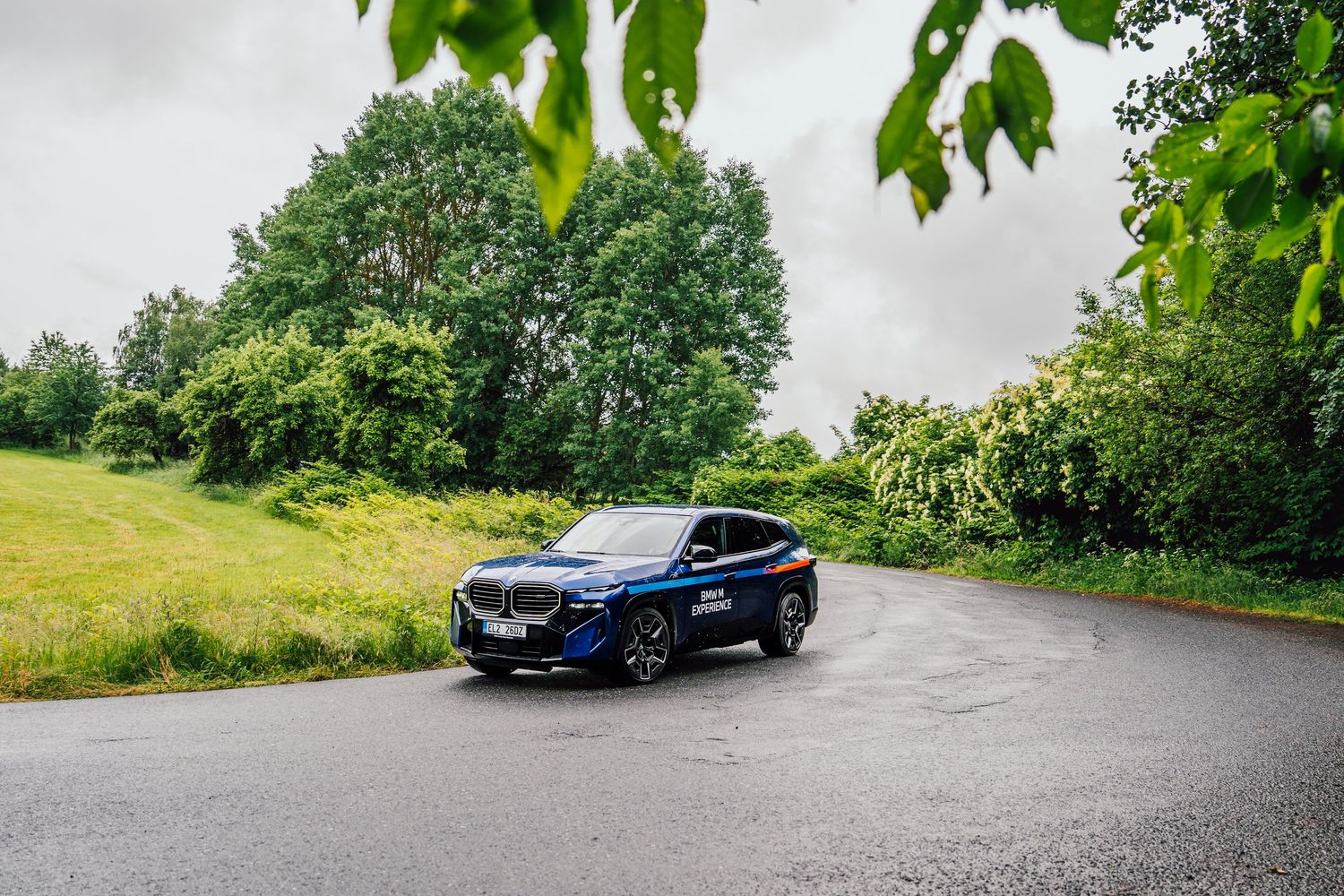 Blue BMW M Experience SUV parked on a quiet road surrounded by green countryside landscape.