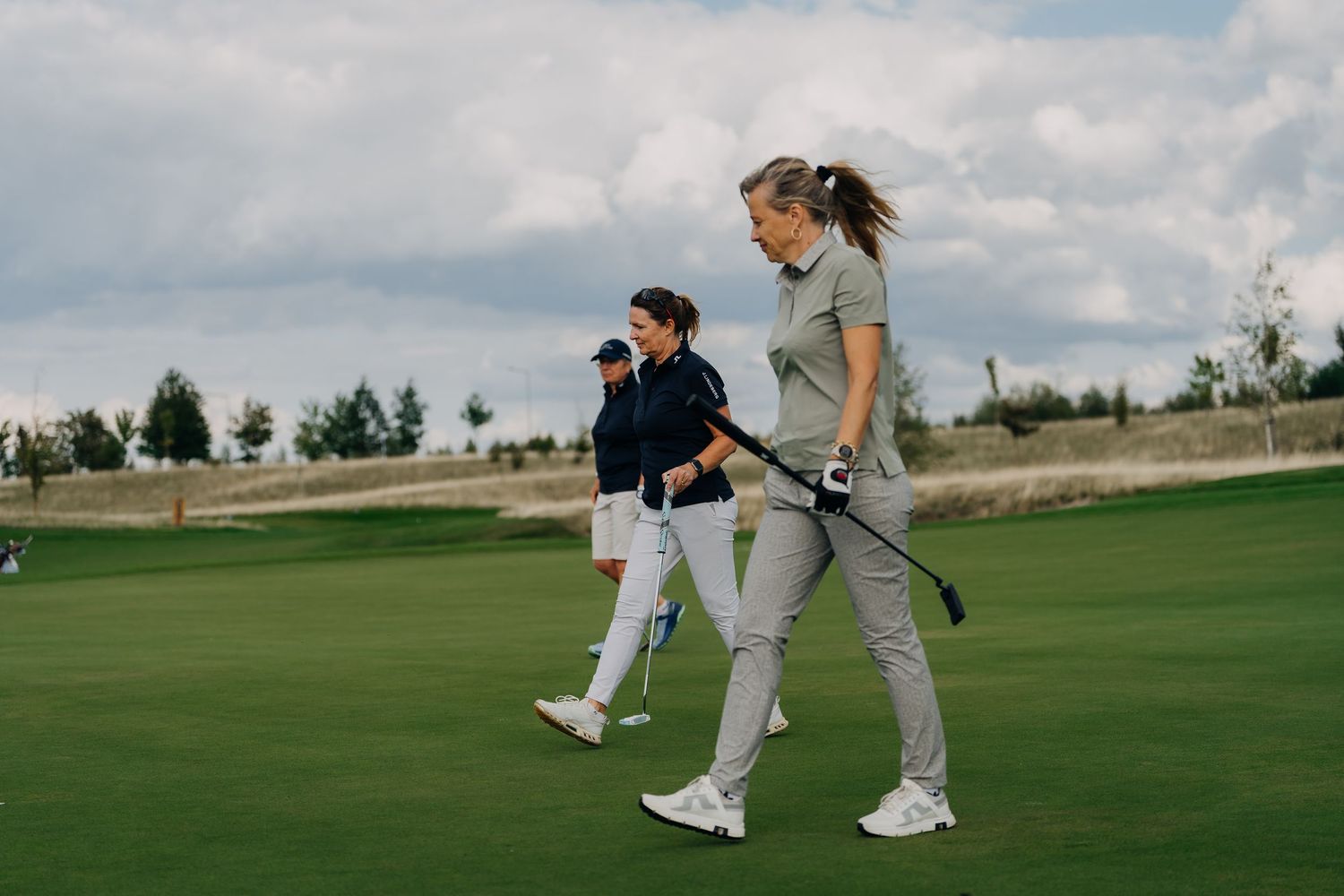 Three golfers walking on a lush green golf course during a round, woman in foreground holding a club.