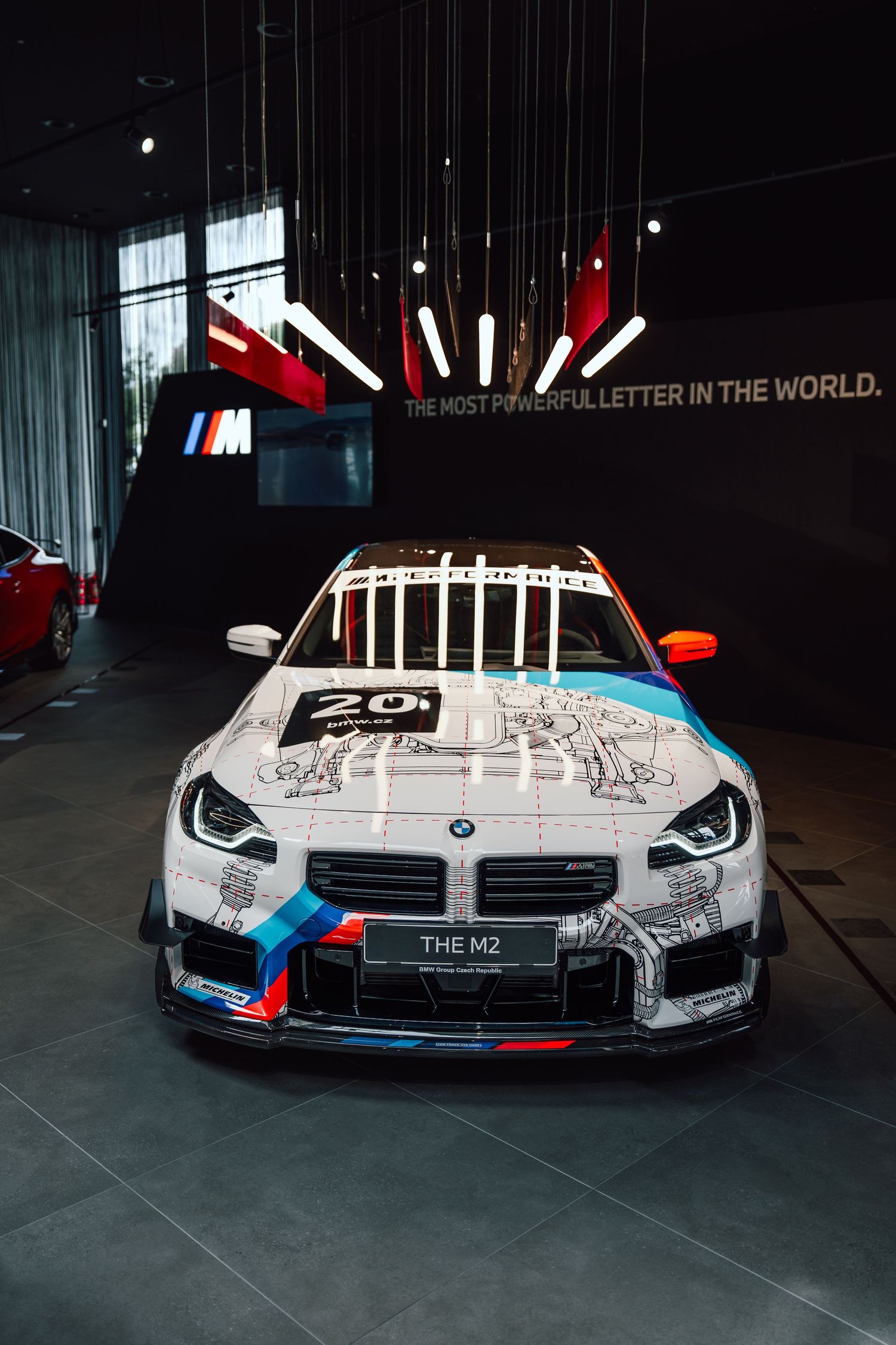BMW M2 race car number 20 with blueprint livery displayed indoors under dramatic lighting at a BMW showroom.