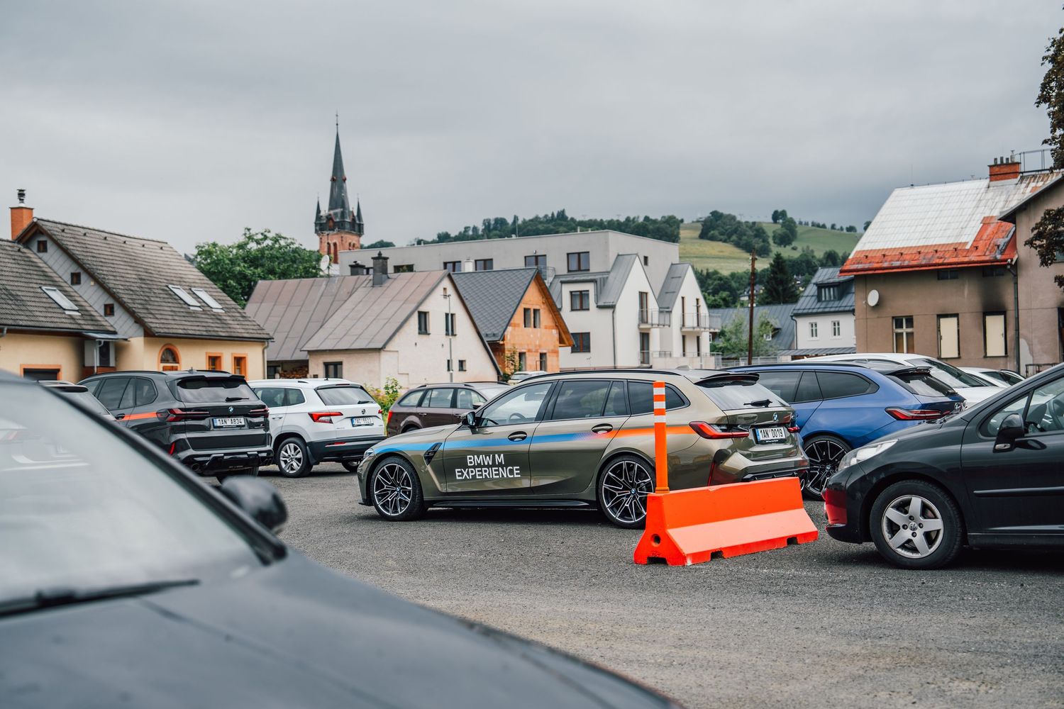 BMW M Experience cars gathered in a town square parking lot with a European church in background.