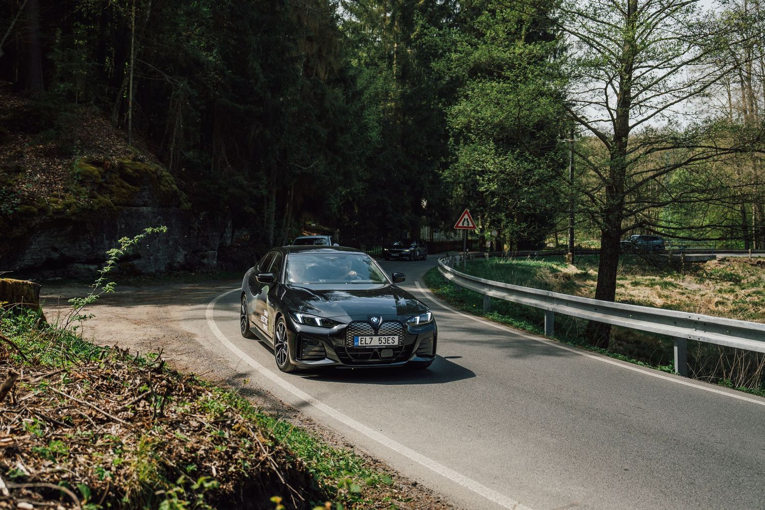 Black BMW sedan driving on a winding road through a sunlit forest with green trees on both sides.