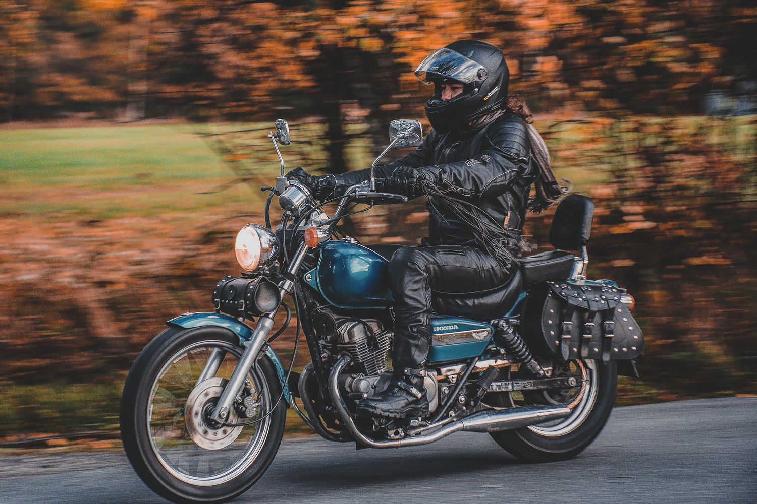 Biker in full leather gear riding classic blue cruiser motorcycle on autumn road with motion blur.