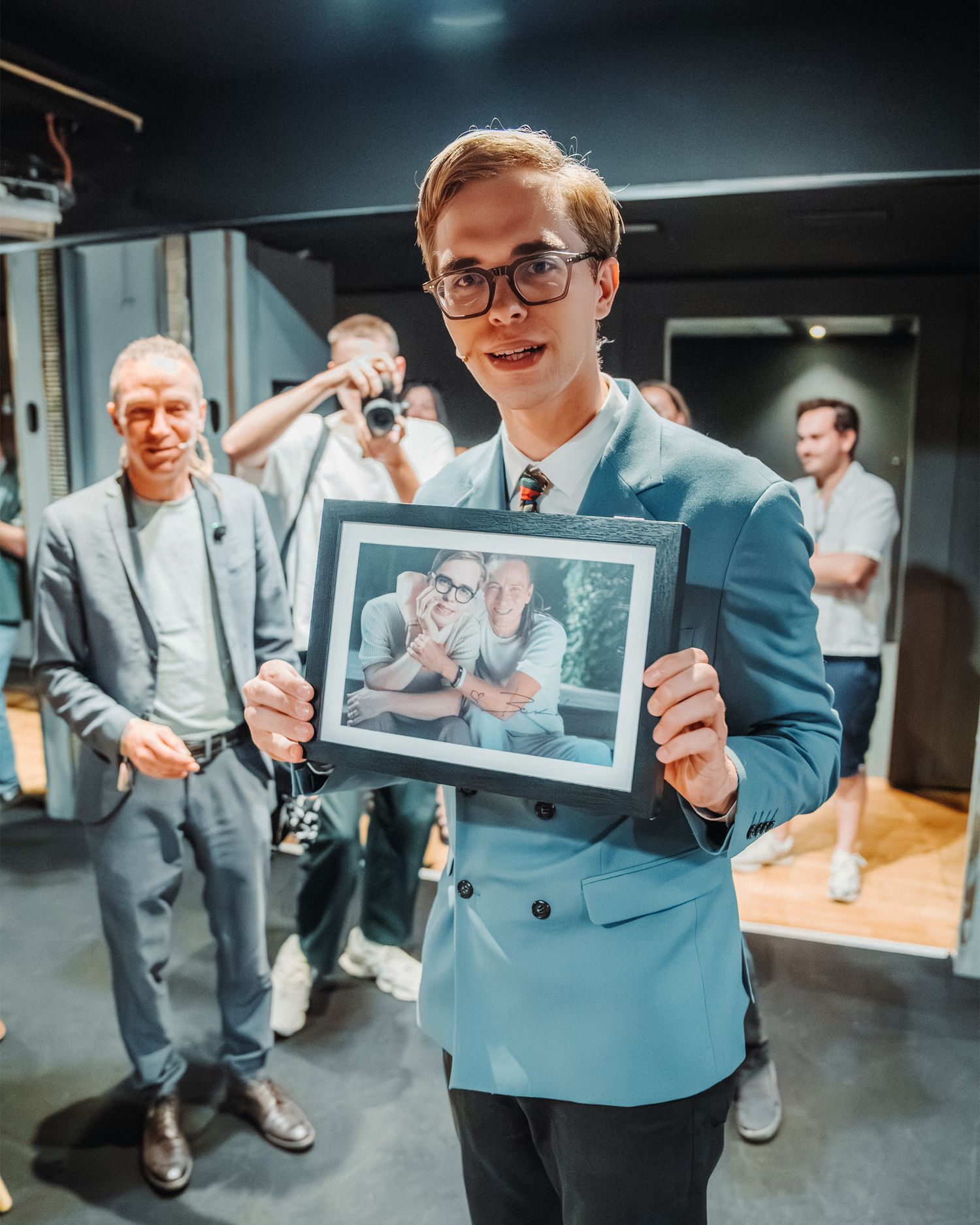 Young man in light blue suit holding a framed photo at an indoor event with people in background.