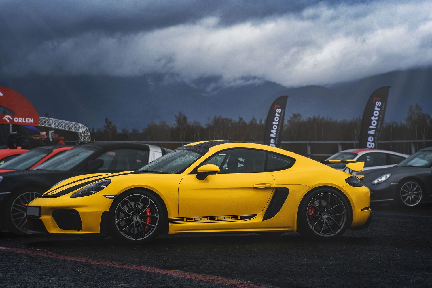 Yellow Porsche 718 Cayman GT4 parked at a motorsport event under dramatic stormy skies.